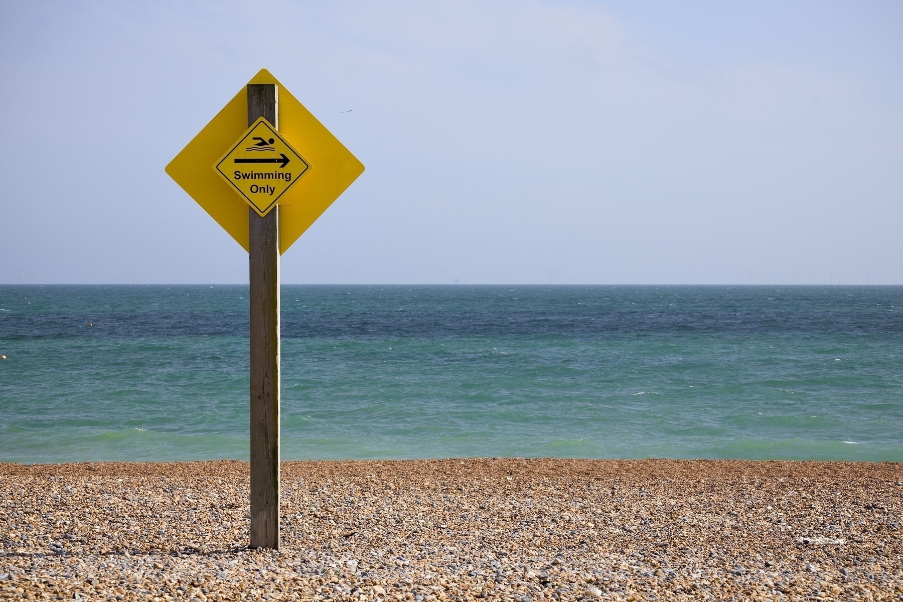 A "Swimming Only" sign stands on a pebbled beach with the sea in the background.