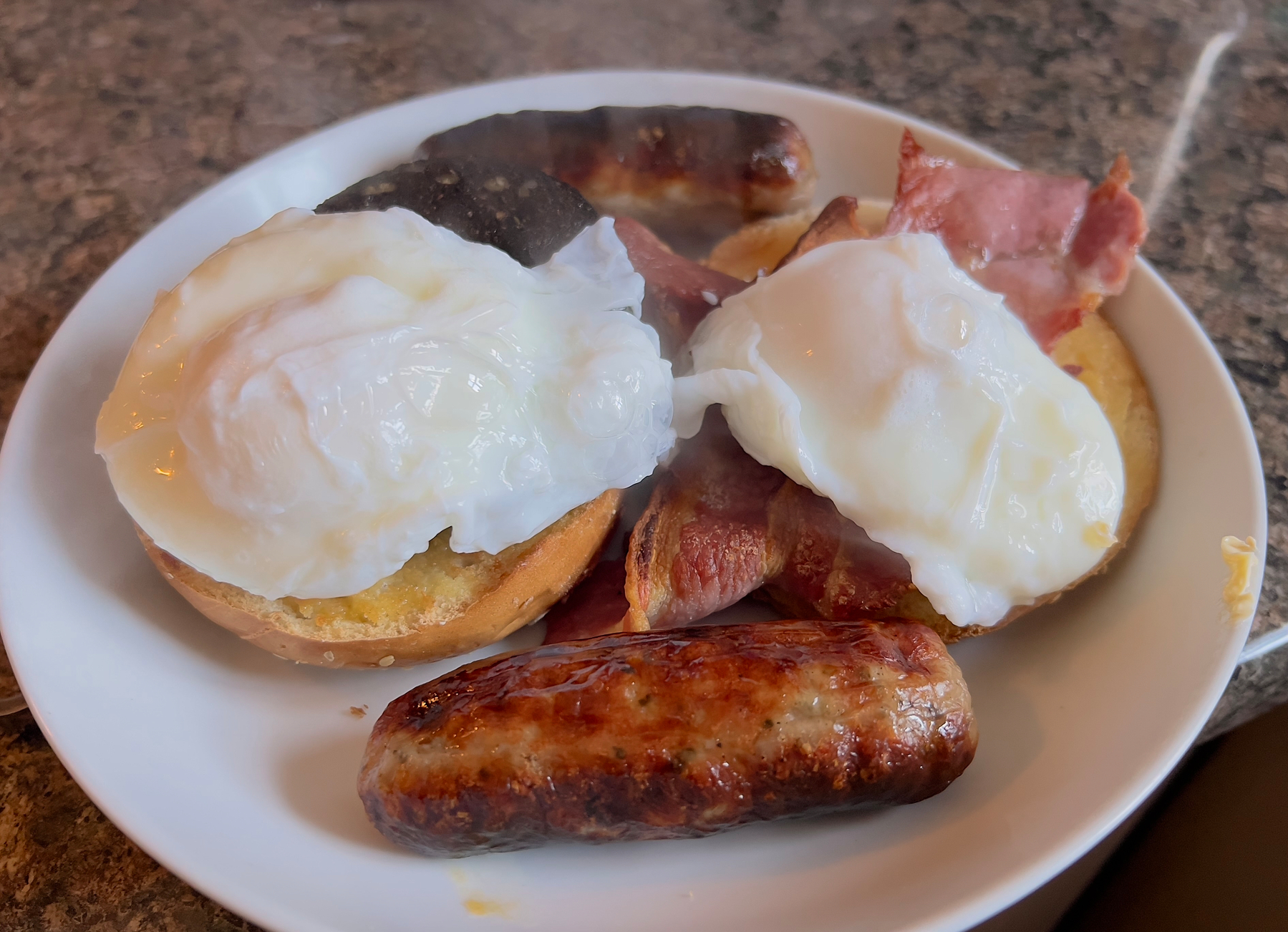 A plate containing poached eggs, sausages, bacon, and a toasted bagel.