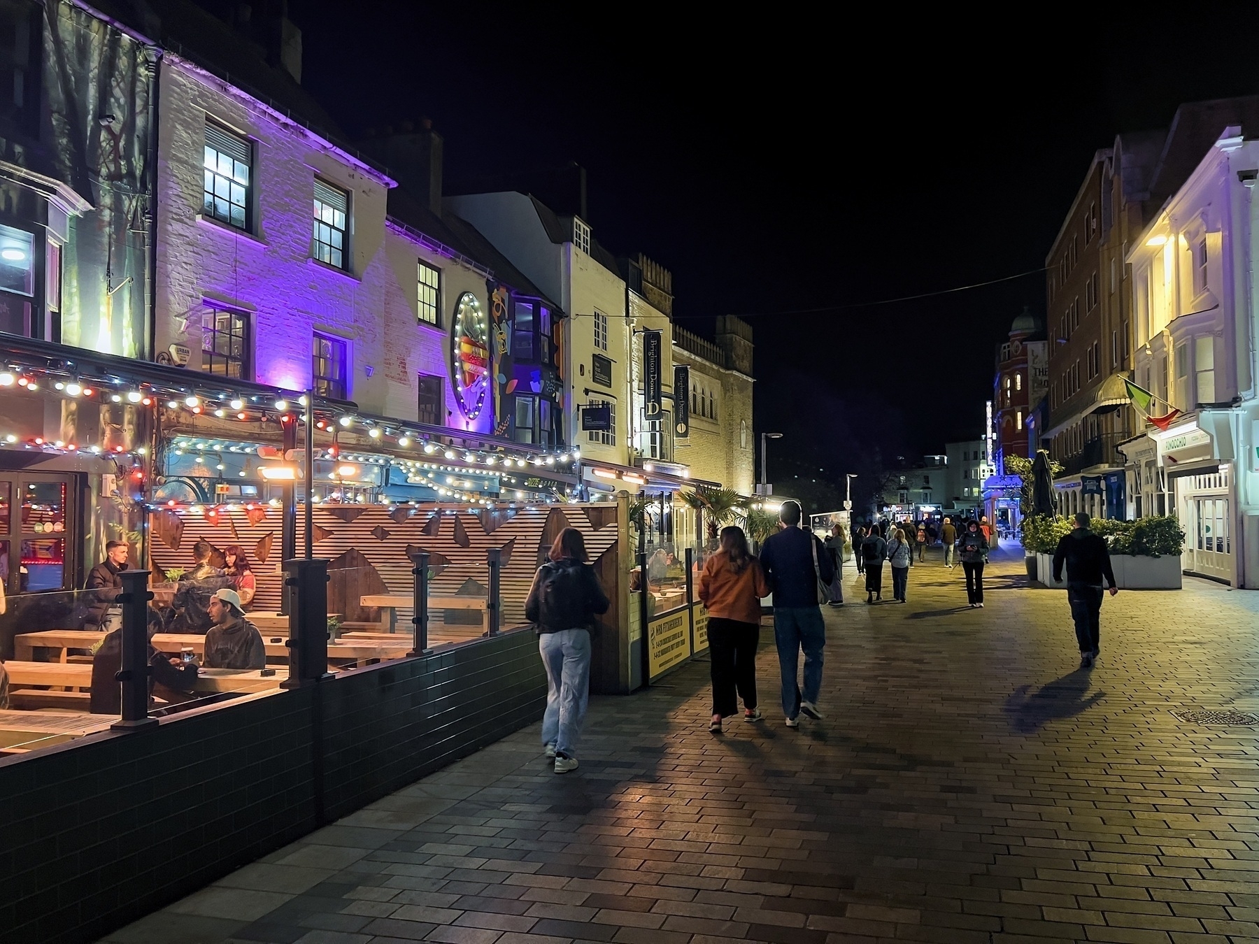 A lively pedestrian street at night with people walking and dining outdoors. Buildings are lit with colorful lights, and a restaurant with string lights has patrons seated inside. The atmosphere is vibrant and festive.