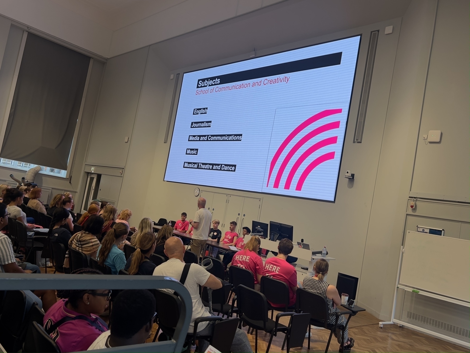 A classroom full of potential students is attending a lecture about subjects in the School of Communication and Creativity, as displayed on a large screen.