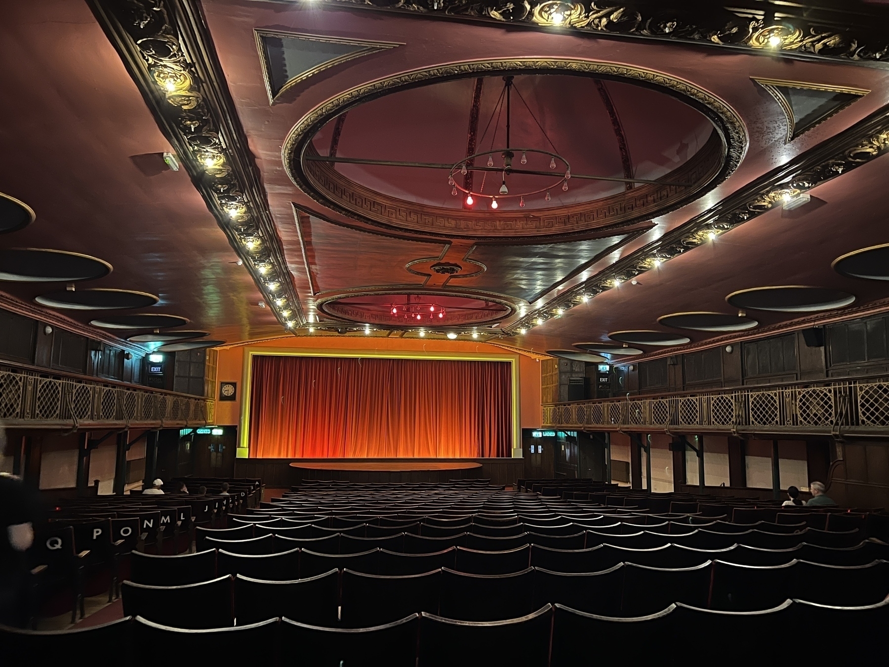 A grand theater with an ornate ceiling and rows of empty seats faces a stage with a red curtain.