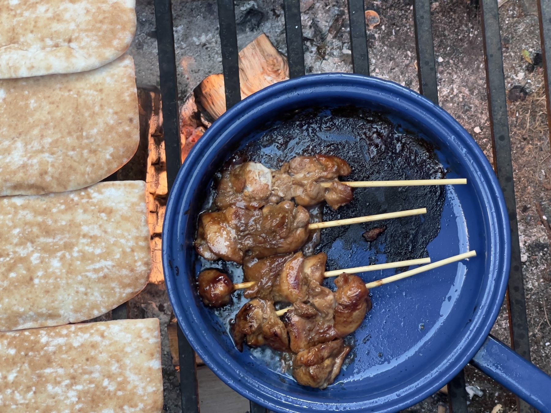 Skewered meat is being grilled in a blue pan over an open flame, accompanied by slices of flatbread.