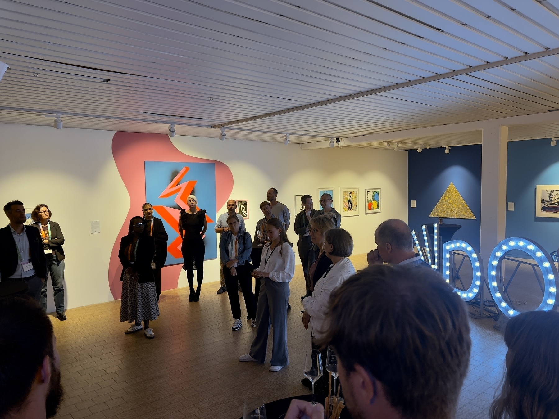 A group of people are gathered in an art gallery, observing a wall with colorful artworks and a neon "DOOMED" sign.