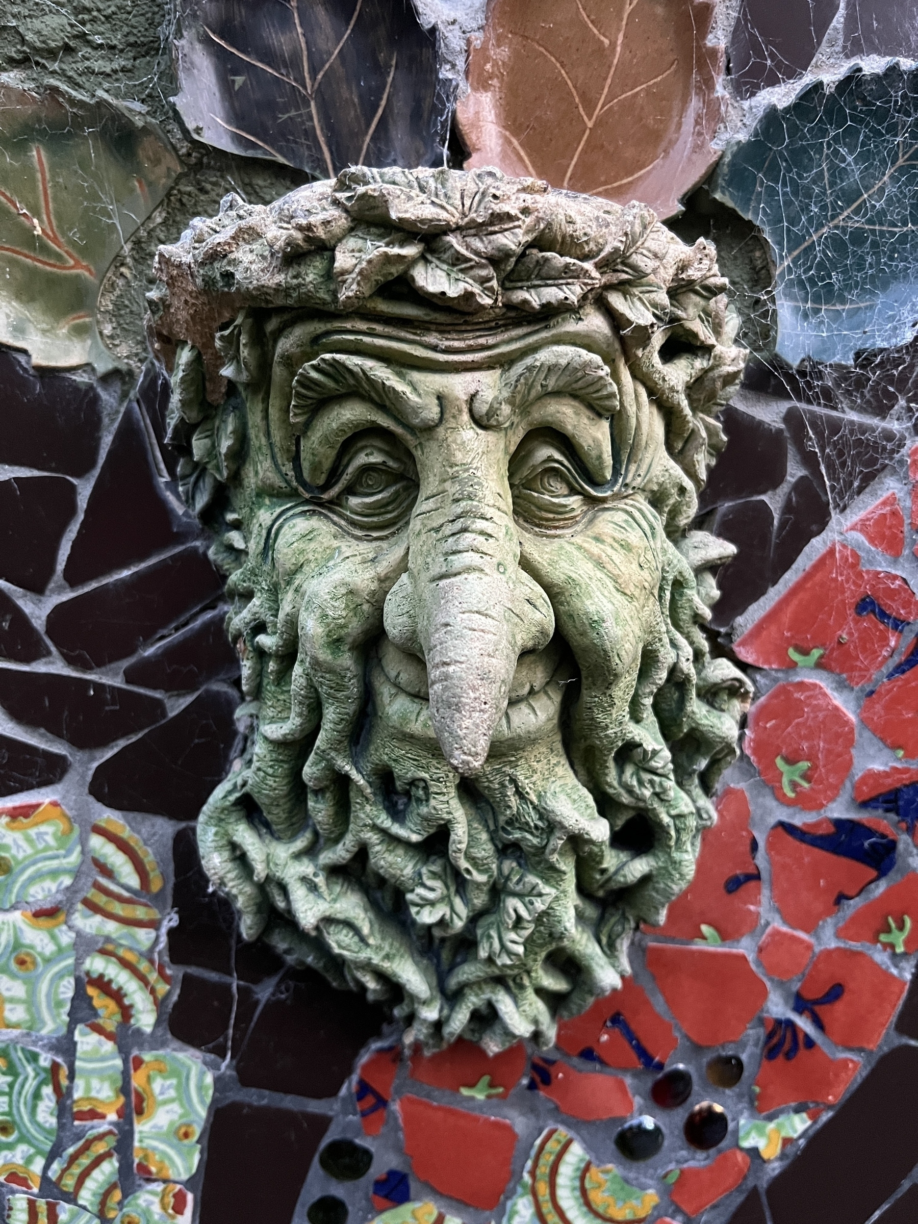 A green, carved face resembling a bearded man with a long nose is surrounded by colorful mosaic tiles and cobwebs.