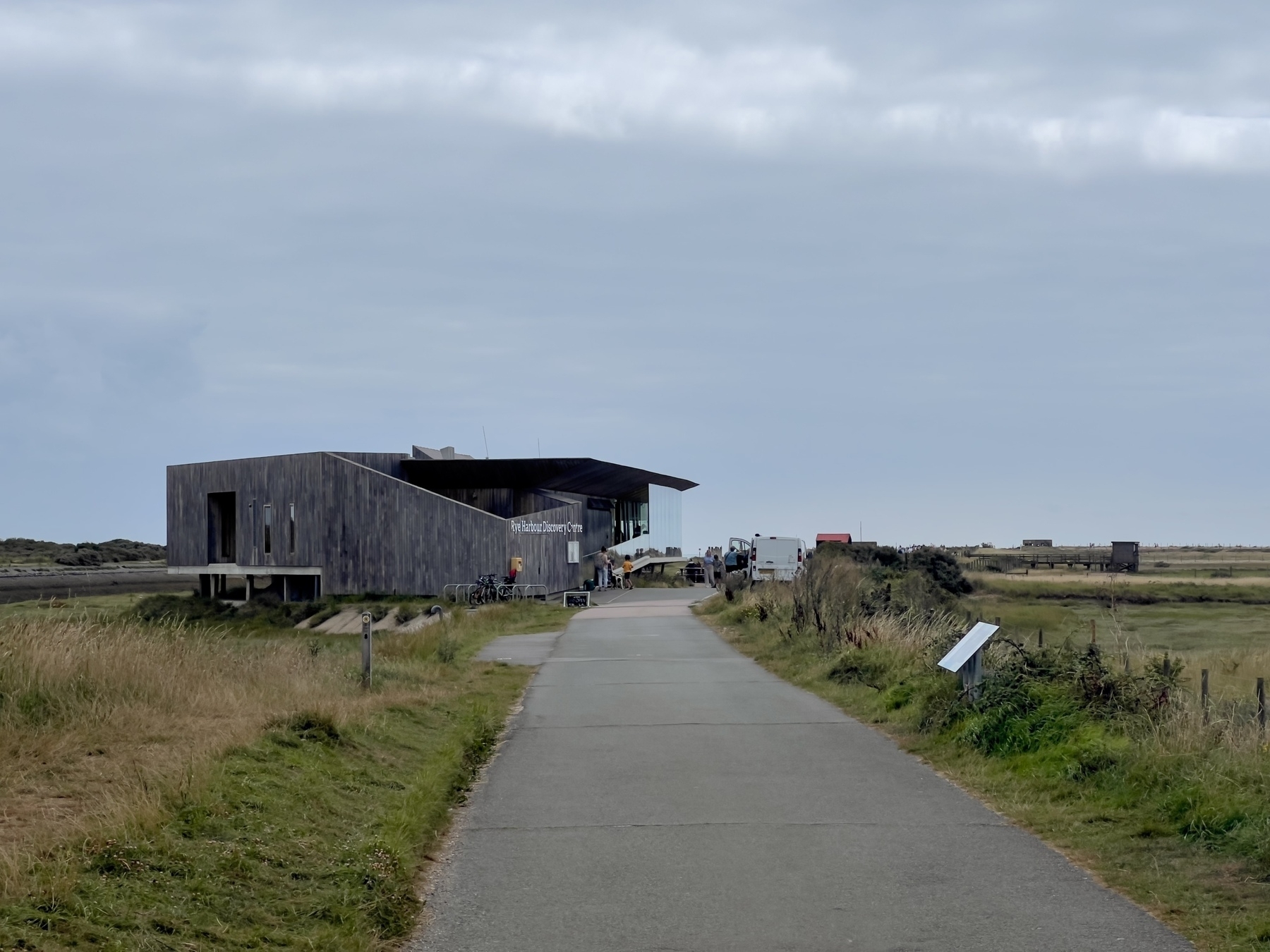 A modern wooden building - the Rye Harbour Discovery  Center - is situated along a pathway in a grassy, rural landscape under a cloudy sky.