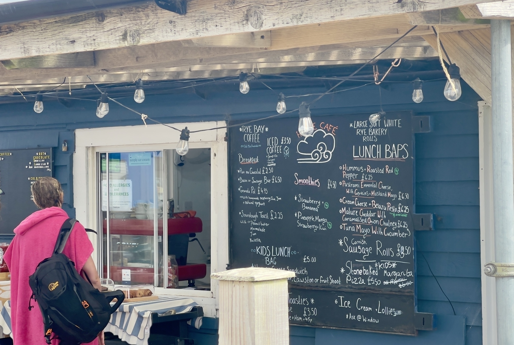 A person in a pink garment stands near a café with a menu board and string lights.