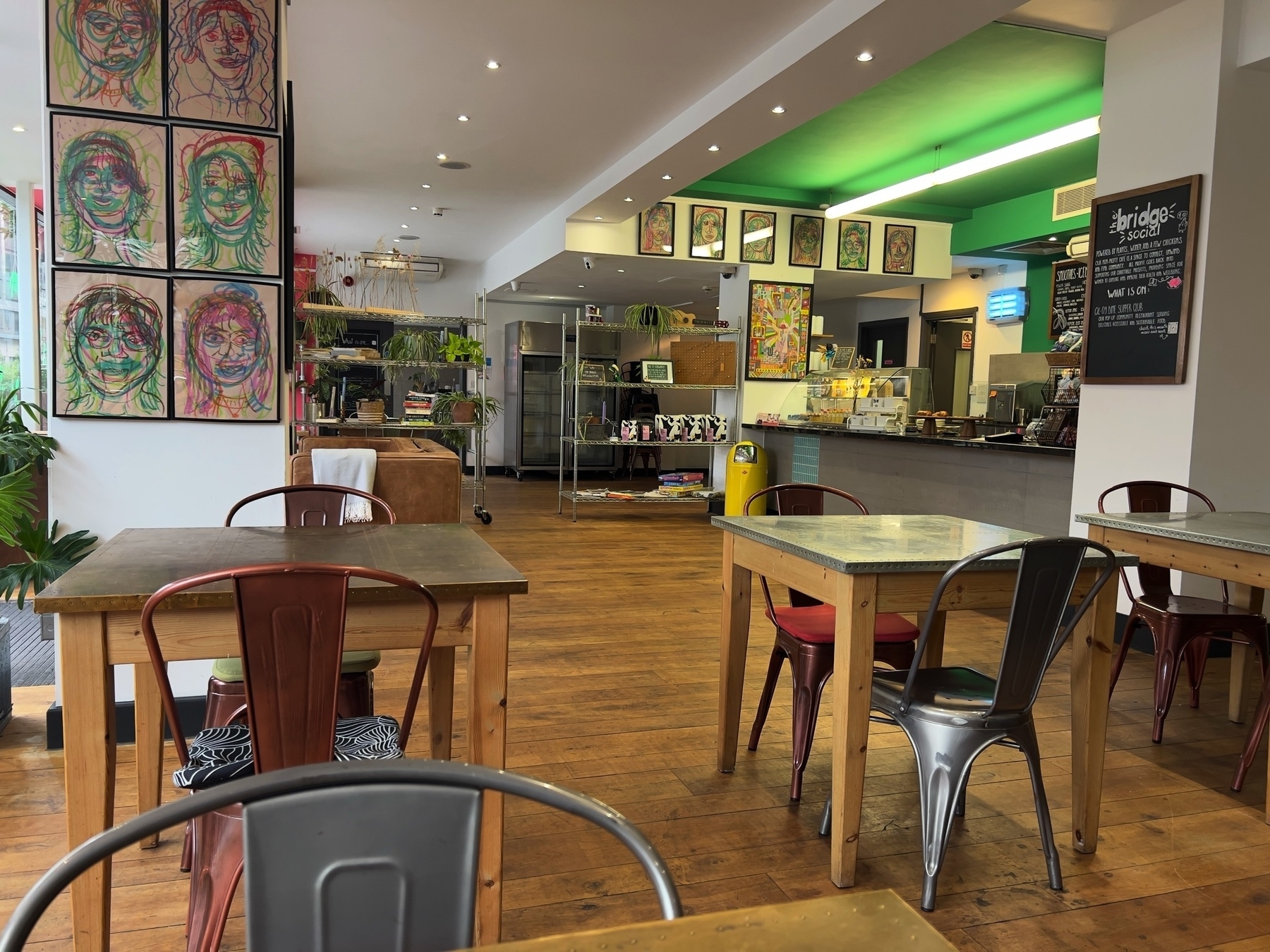 A cozy café features eclectic artwork on the walls, mismatched chairs around wooden tables, and a counter displaying a menu.