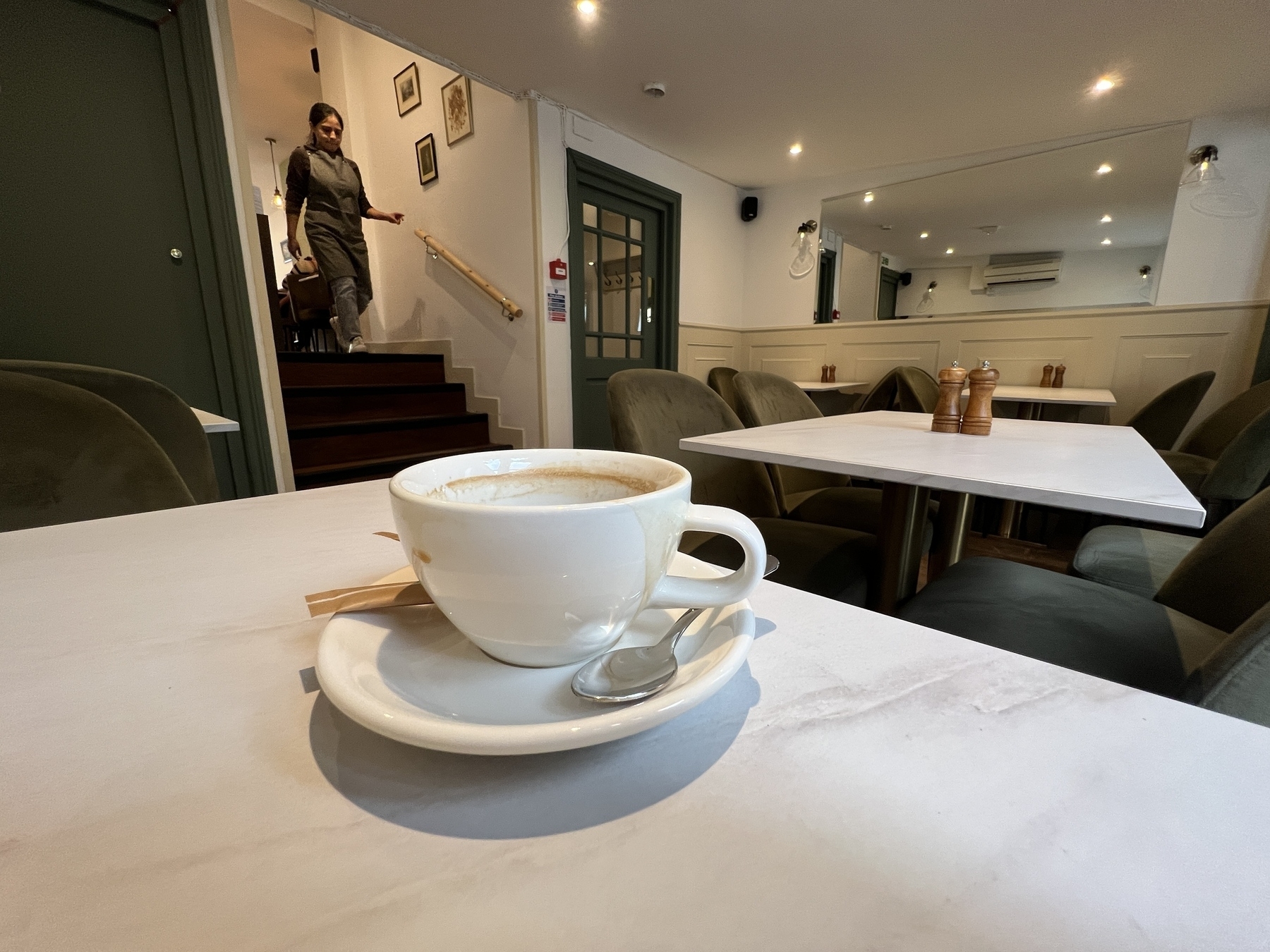 A cup of coffee sits on a table in a modern cafe with a person descending a staircase in the background.