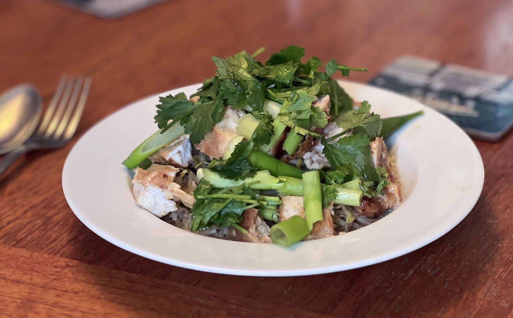 A plate of grilled chicken salad topped with fresh greens and herbs is served on a wooden table.