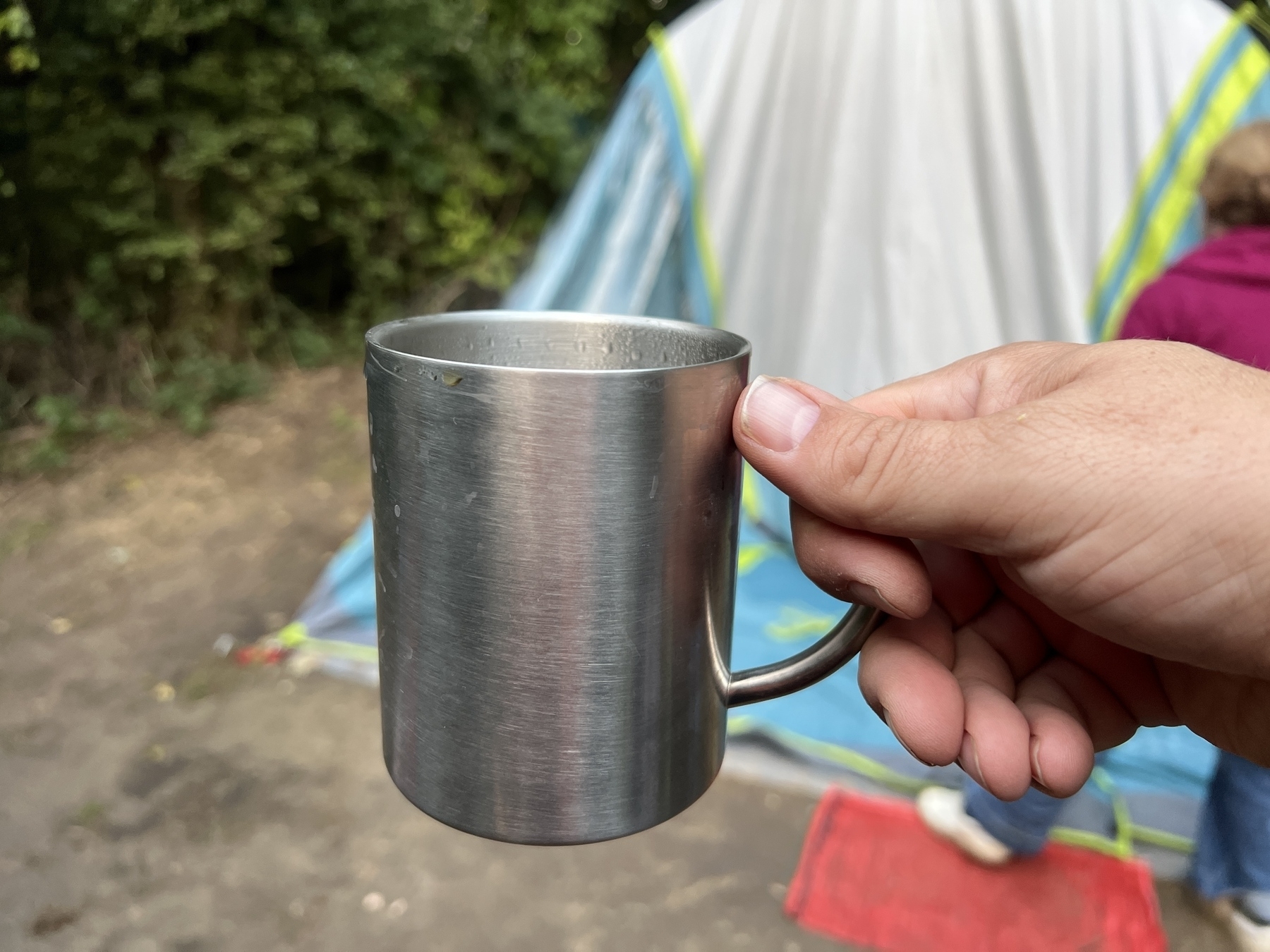 A hand is holding a stainless steel mug in front of a tent outdoors.