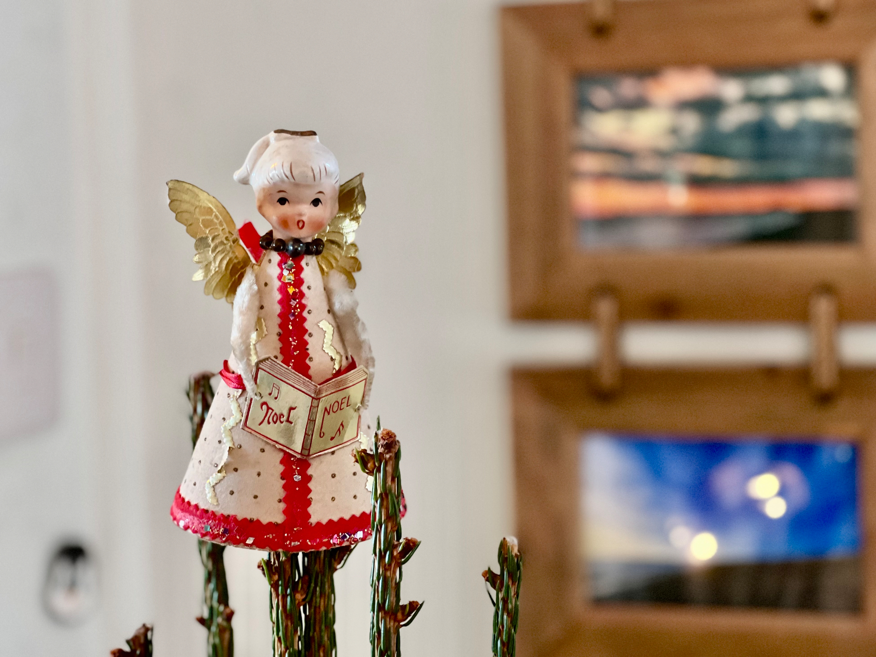 A decorative figurine of an angel holding a book with Joyeux Noël written on it stands on top of a Christmas tree in front of a blurred background with framed pictures.