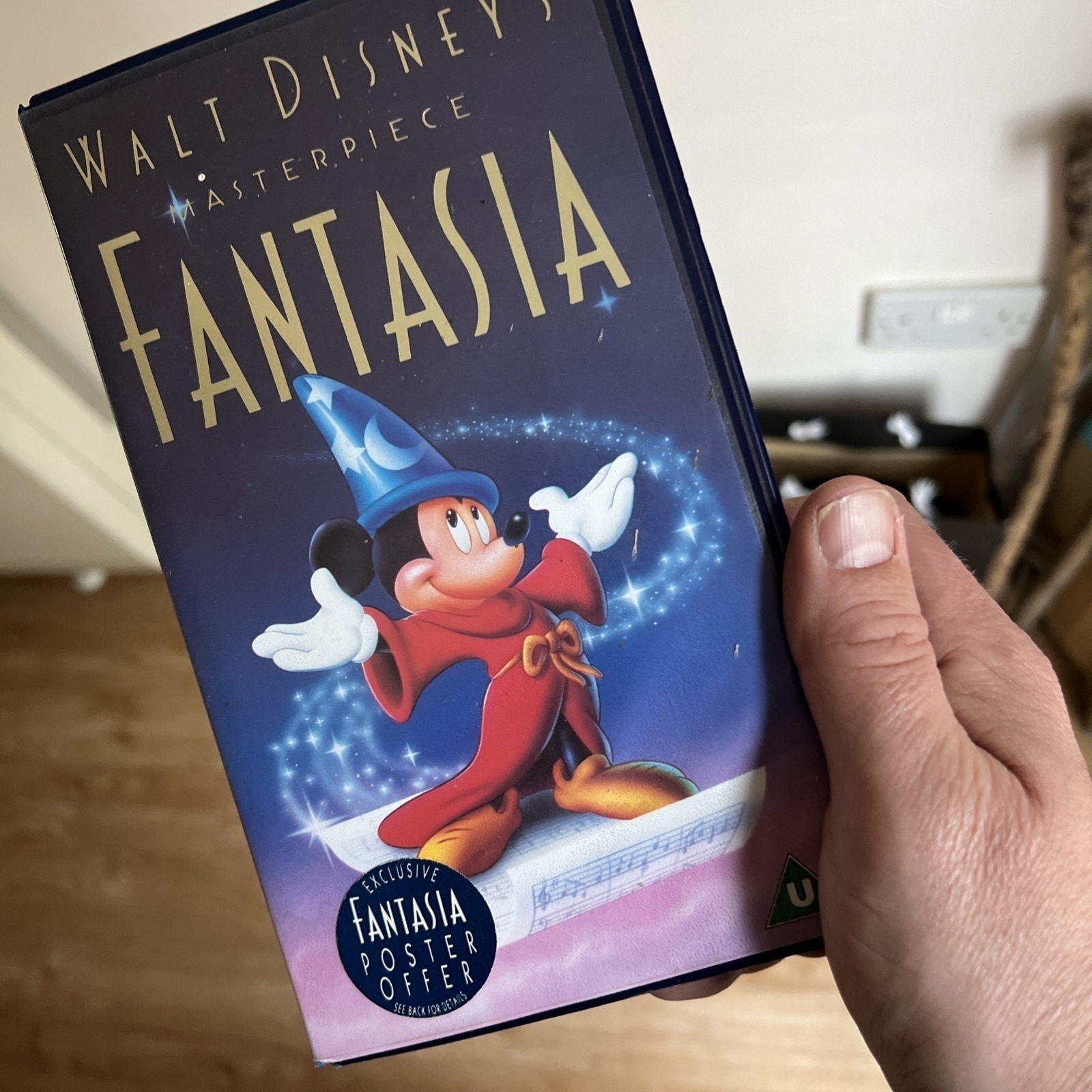 A person is holding a VHS tape of Walt Disney’s animated film “Fantasia.” The cover features Mickey Mouse dressed as the Sorcerer’s Apprentice, wearing a blue wizard hat and red robe, surrounded by magical sparkles. The box also advertises an exclusive Fantasia poster offer.