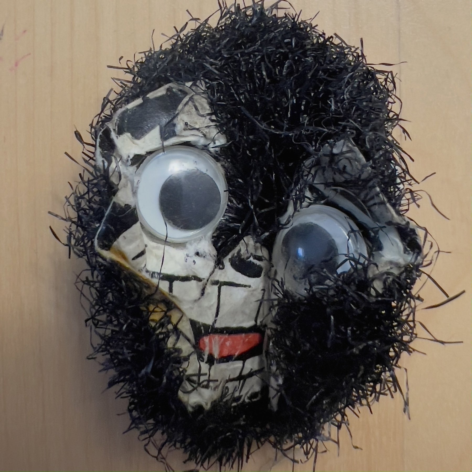 A badge with googly eyes, a black and white face, and black hair-like material on a wooden background.
