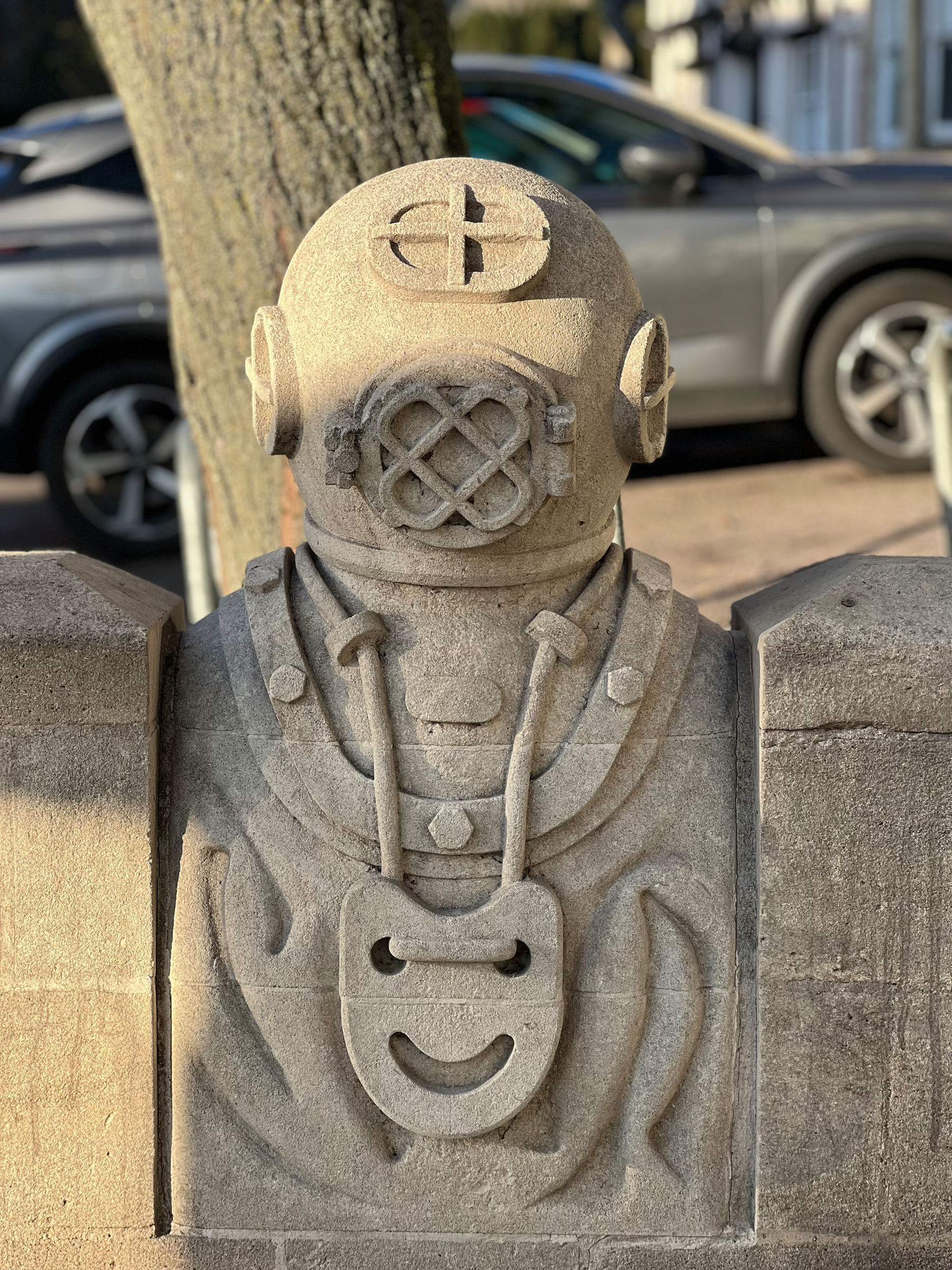 A stone sculpture of an old-fashioned diving helmet is mounted on a concrete base, situated near a tree and parked car.