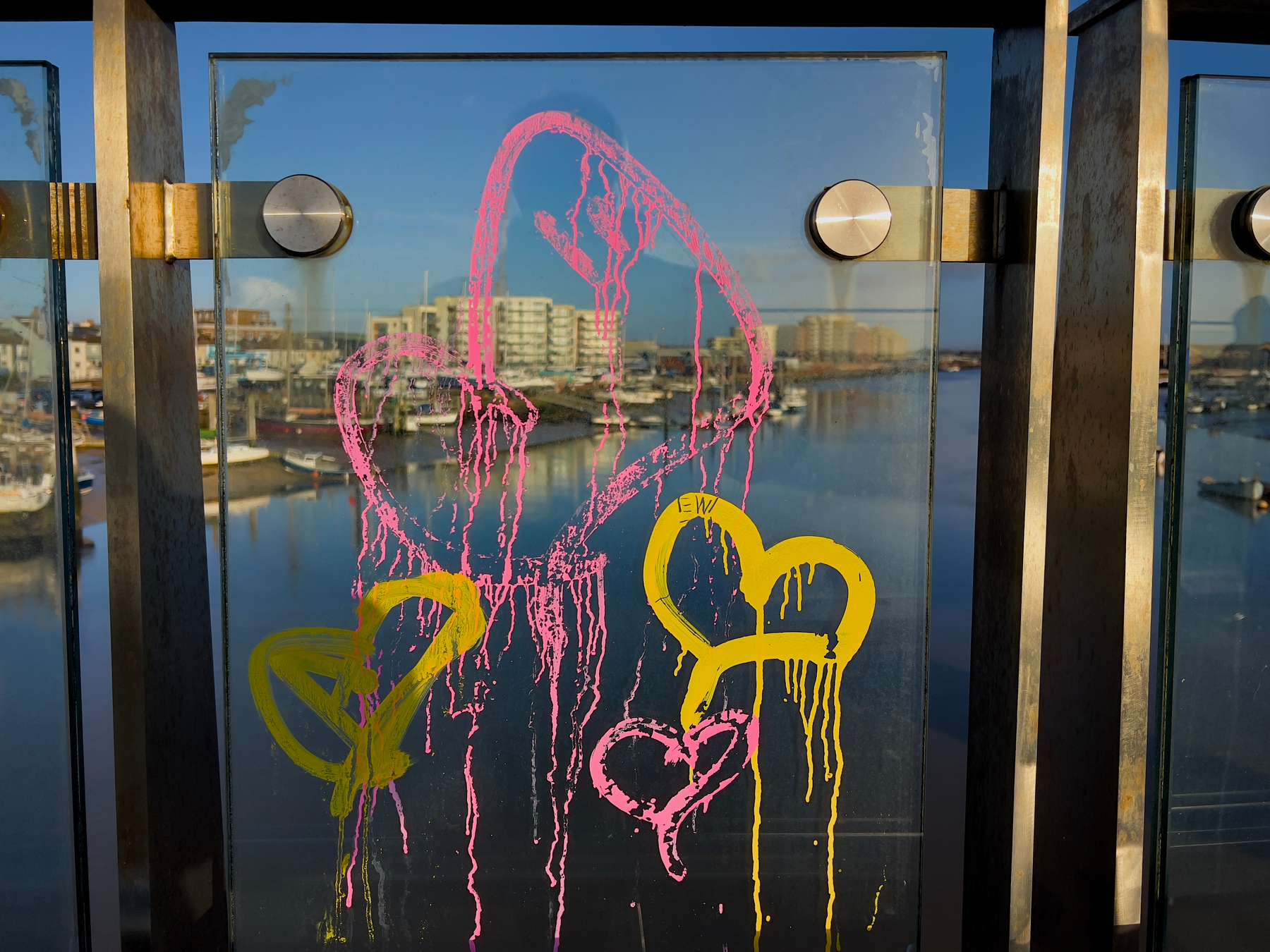 Brightly colored hearts, drawn in pink and yellow paint, decorate a glass panel overlooking a waterfront with buildings and boats.