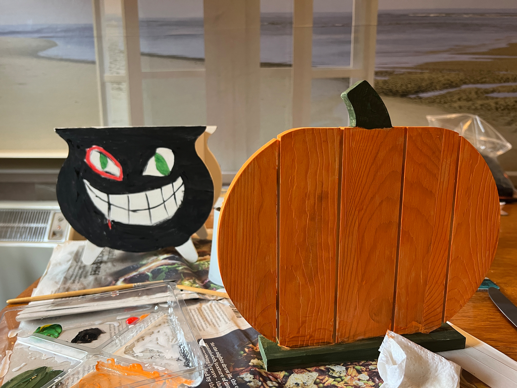 A painted wooden cauldron with a grinning face and a wooden pumpkin are displayed on a table with paint supplies.