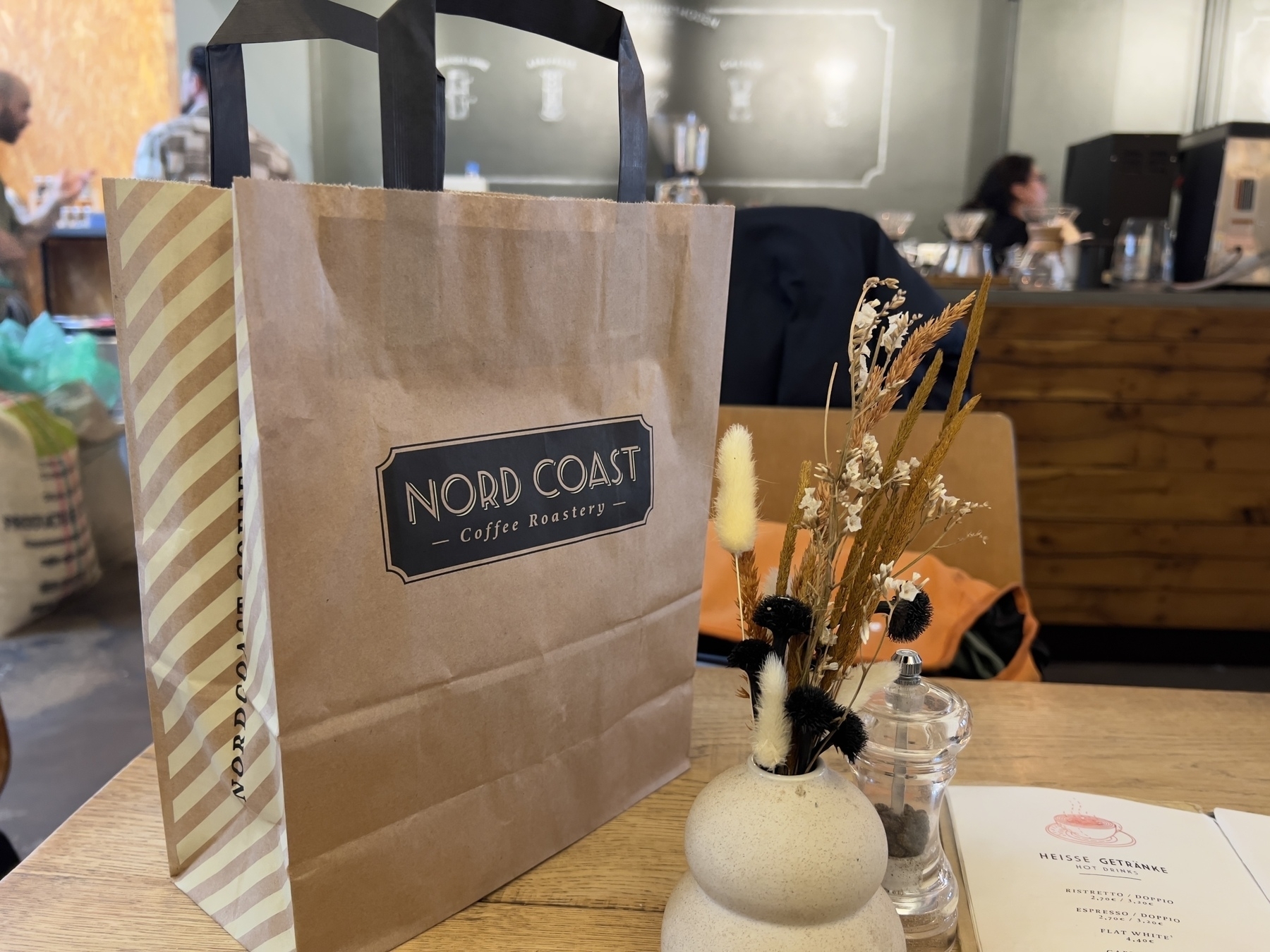 A brown paper bag from "Nord Coast Coffee Roastery" sits on a table next to a small vase with dried flowers in a cozy café setting.