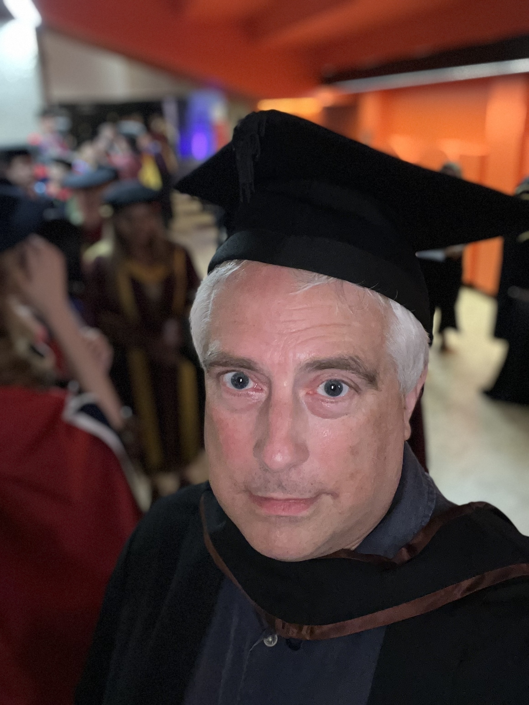 A person wearing a graduation cap and gown is taking a selfie in a brightly colored indoor setting.
