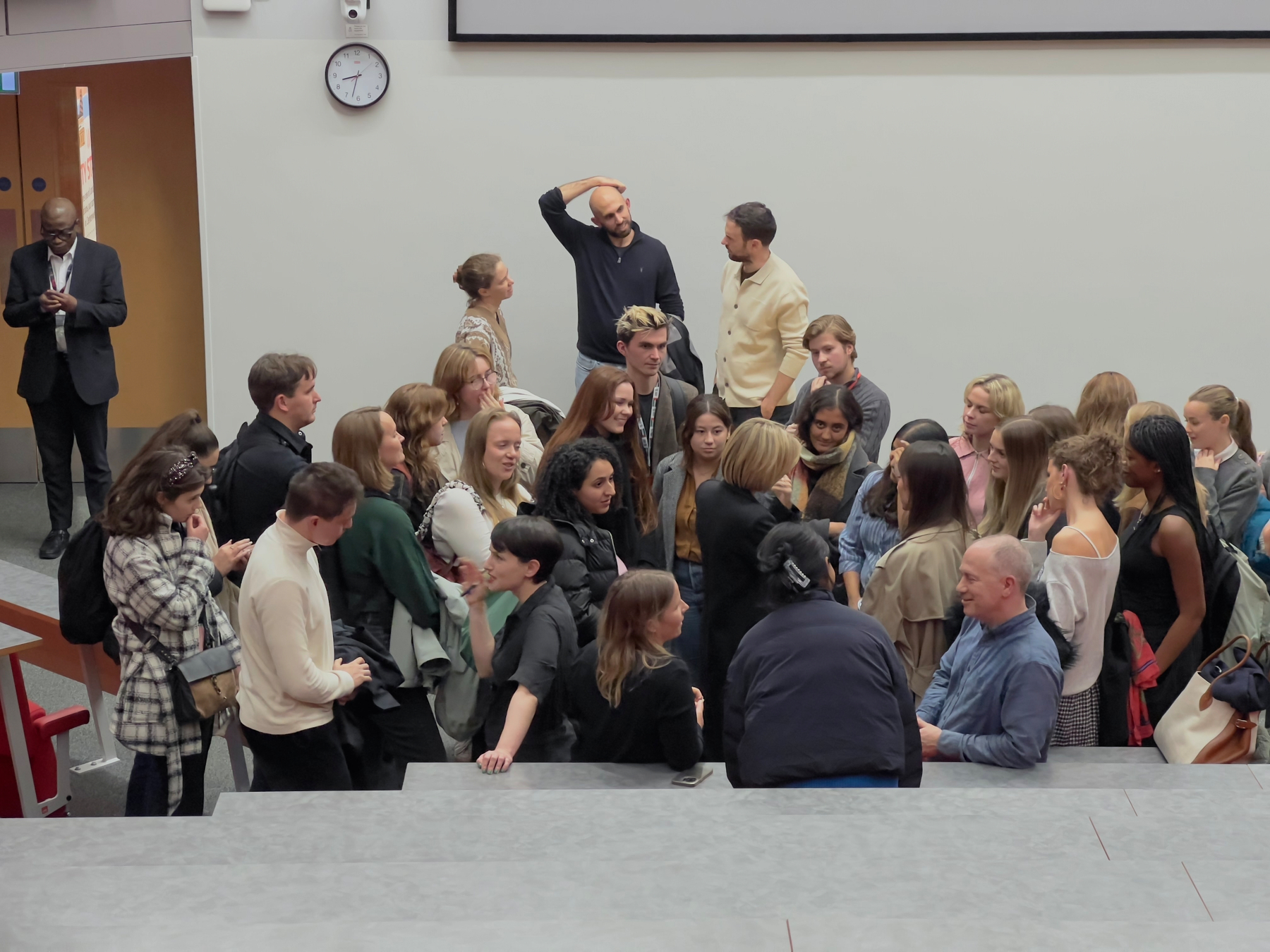 A group of people is gathered in an auditorium, engaging in conversations and taking photos.