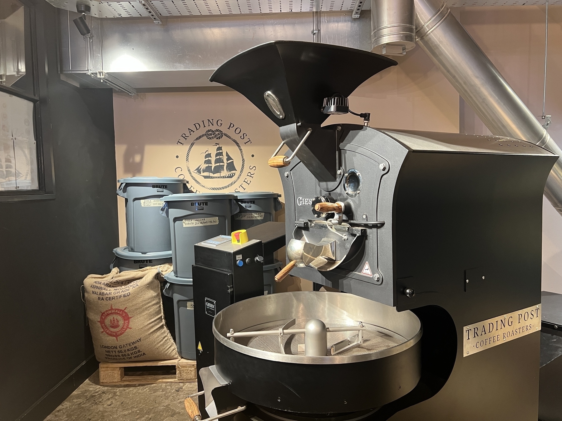 A coffee roasting machine is situated in an industrial setting, surrounded by bags and barrels, with a "Trading Post Coffee Roasters" logo on the wall.