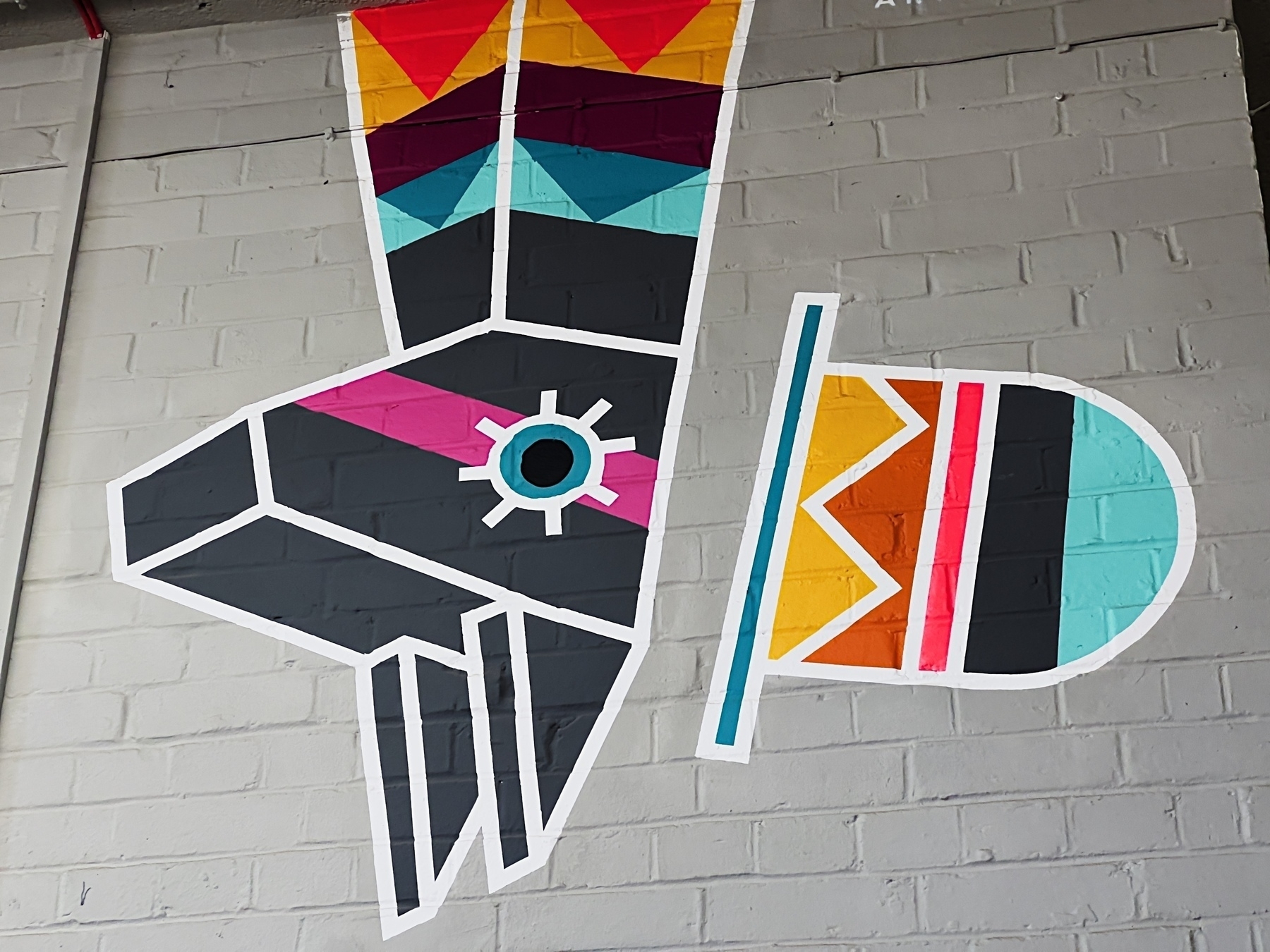 A colorful, geometric mural on a brick wall features abstract shapes resembling a stylised alpaca and hat.