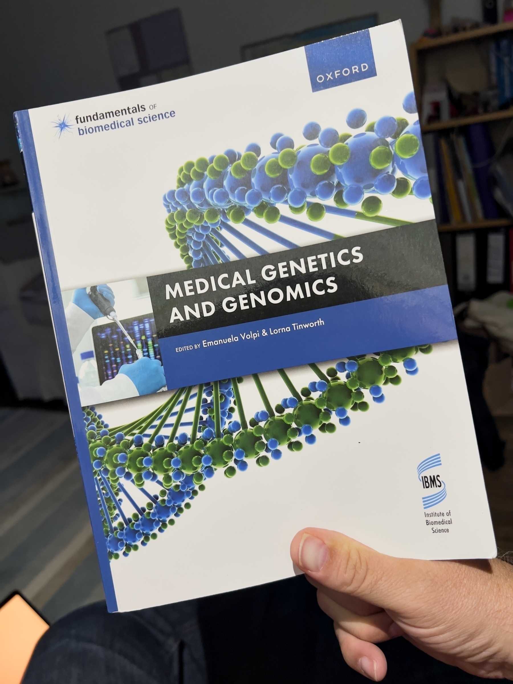 A person is holding a book titled "Medical Genetics and Genomics," part of the Fundamentals of Biomedical Science series from Oxford.