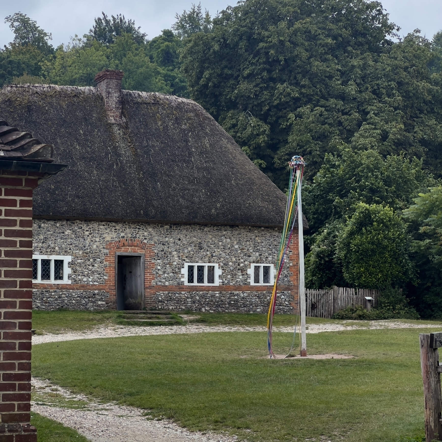 A quaint thatched-roof cottage is surrounded by lush greenery and features a maypole in the yard.