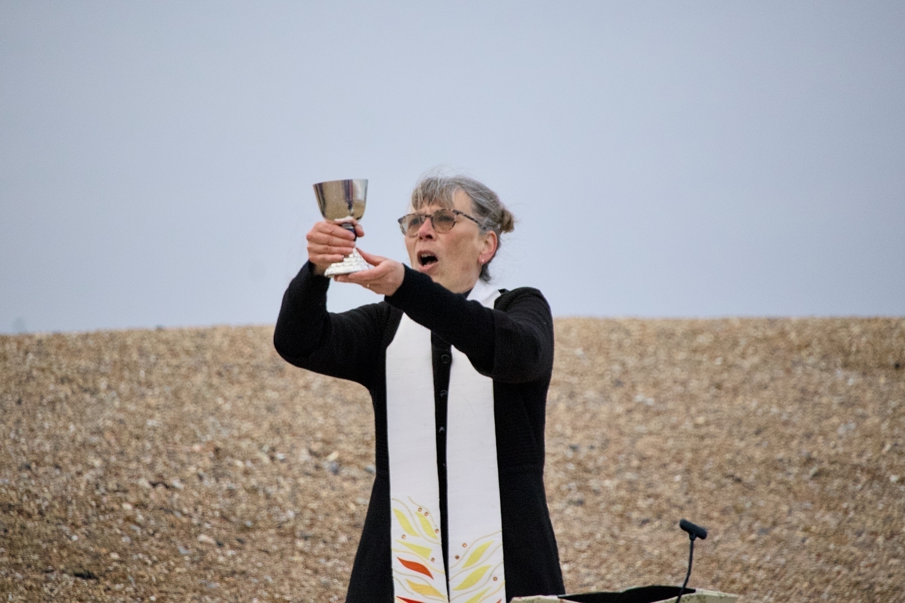A person wearing clerical attire and a stole is holding up a chalice outdoors on a rocky surface.