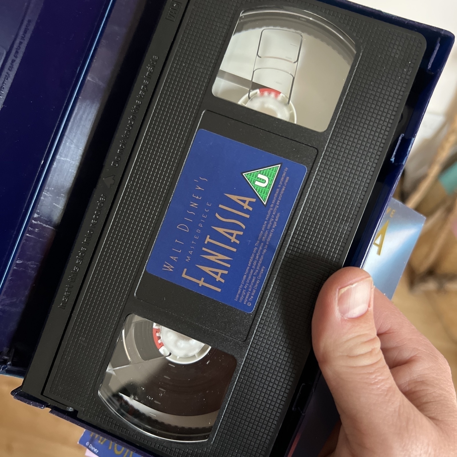 A person is holding a VHS tape of Walt Disney’s animated film “Fantasia” inside its open plastic case. The black cassette features a blue label with gold text reading “Walt Disney’s Masterpiece Fantasia” and a green “U” rating symbol, indicating it is suitable for all audiences in the UK. The tape reels are visible through the clear plastic windows.