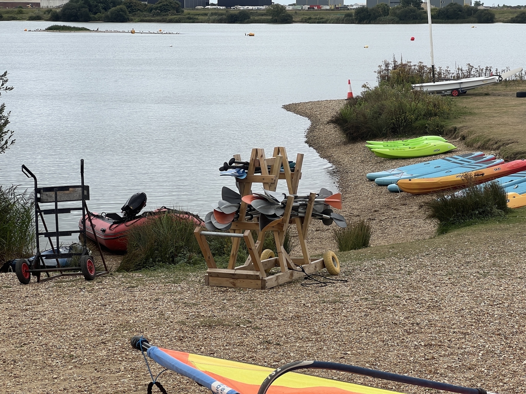 Kayaks and paddles are arranged along a pebbled lakeside shore with a grassy area and a small trailer nearby.