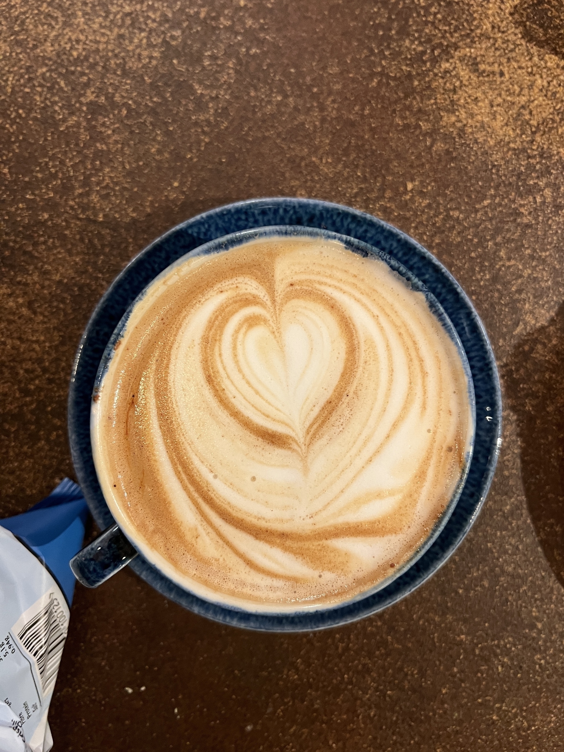 A blue cup holds a latte with a heart-shaped milk foam design on a brown surface.