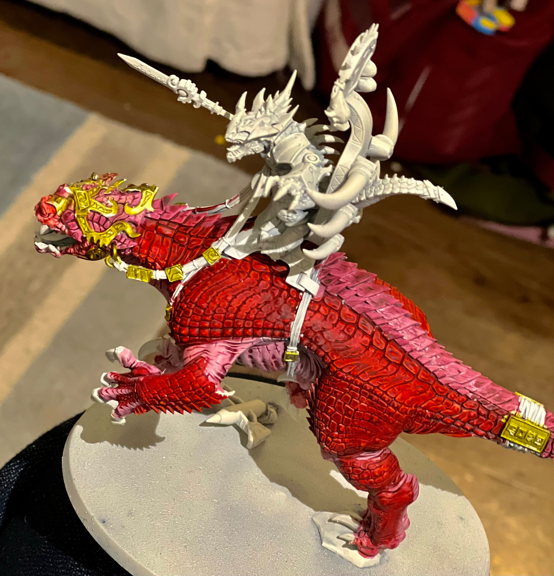 A detailed miniature figure depicts a fantasy creature with red scales and ornate gold accents, carrying an intricately armored rider wielding a spear.