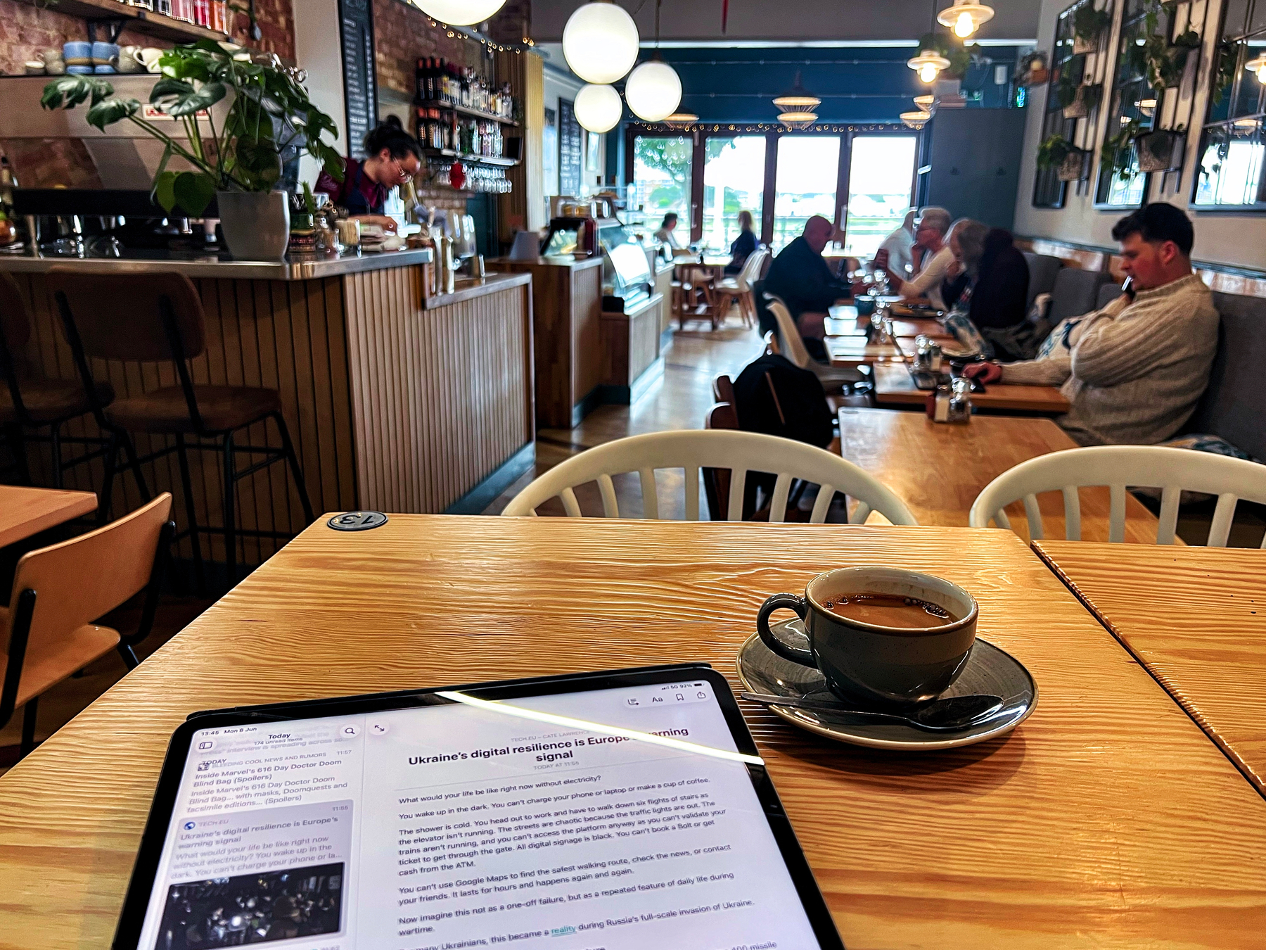 A bland café features several people seated at tables, with one table displaying a tablet and a cup of coffee in the foreground.