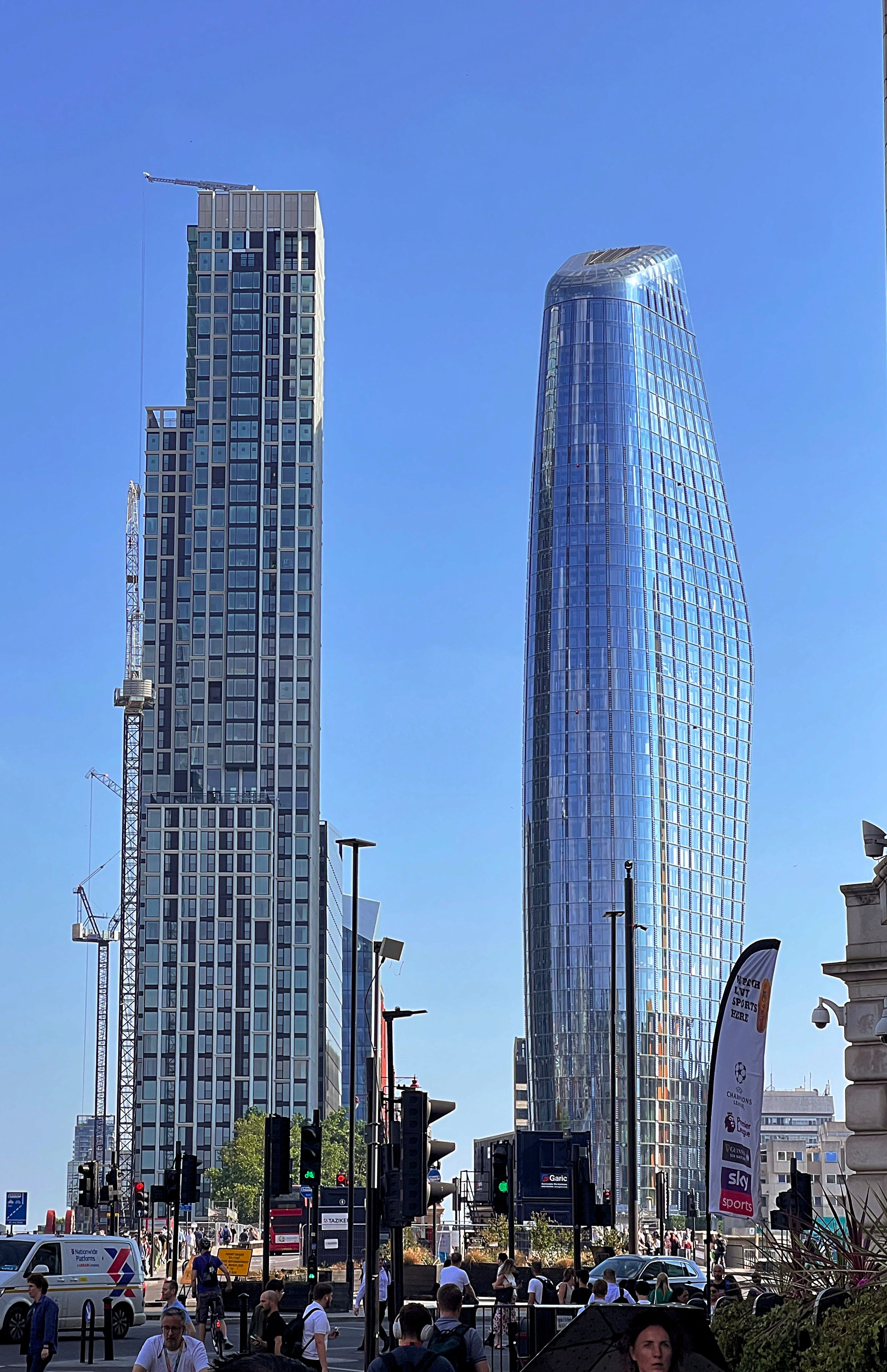 Two distinct modern skyscrapers, one with a rectangular design and the other with a curved, glass facade, stand prominently against a clear blue sky in an urban setting with pedestrians and traffic signals below.