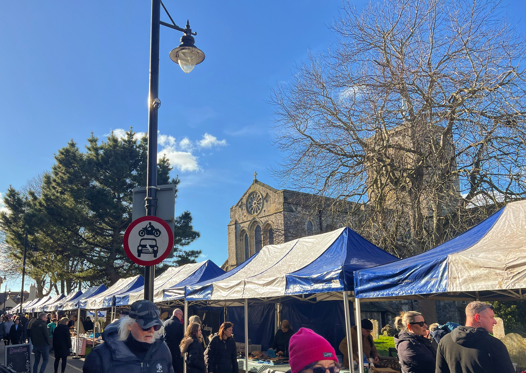 A bustling outdoor market in Shoreham-by-Sea, West Sussex, with people browsing stalls under blue and white canopies, featuring a large church and leafless trees in the background.