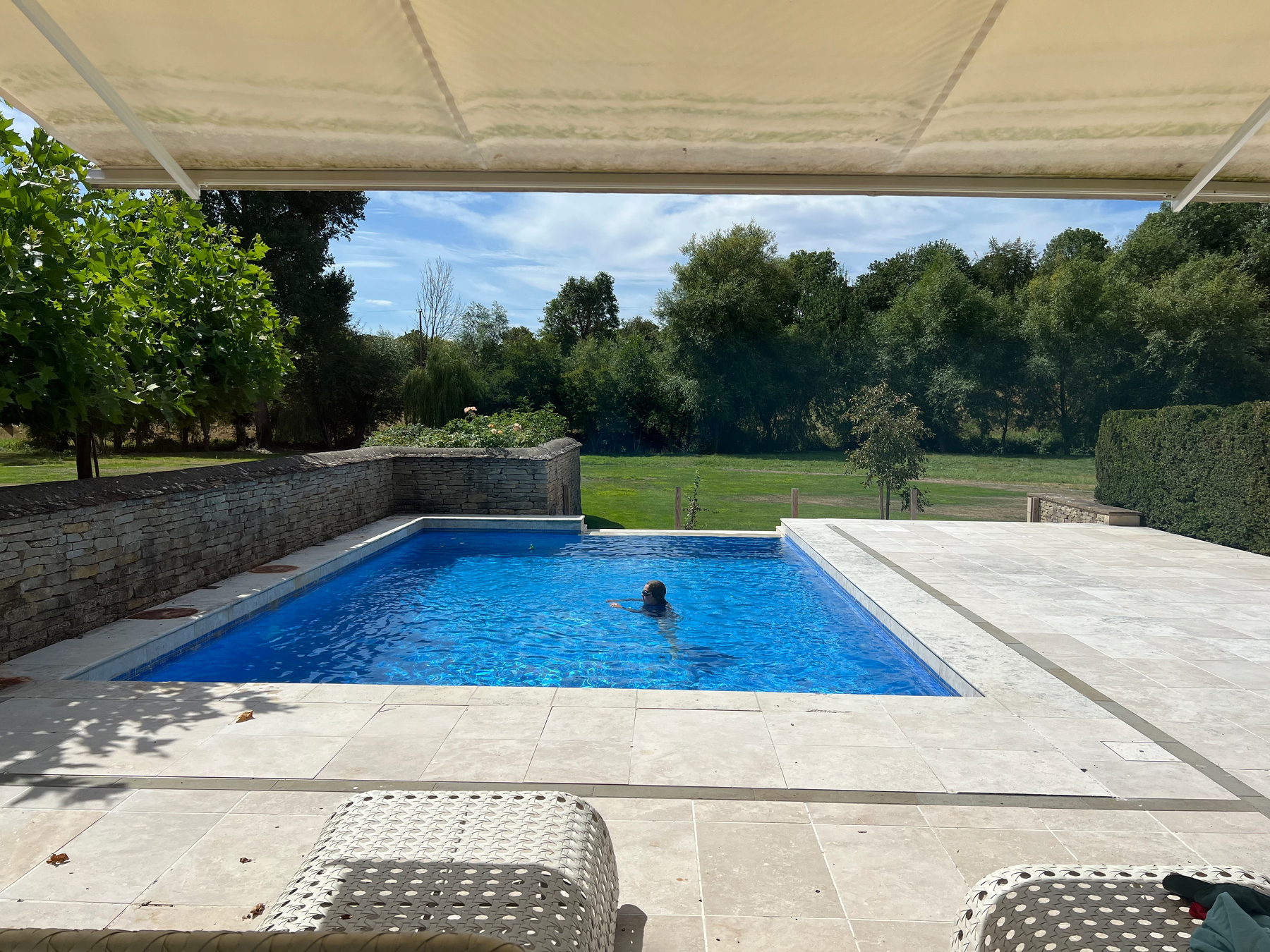 A person is swimming in a rectangular outdoor pool surrounded by a stone patio with greenery and trees in the background.