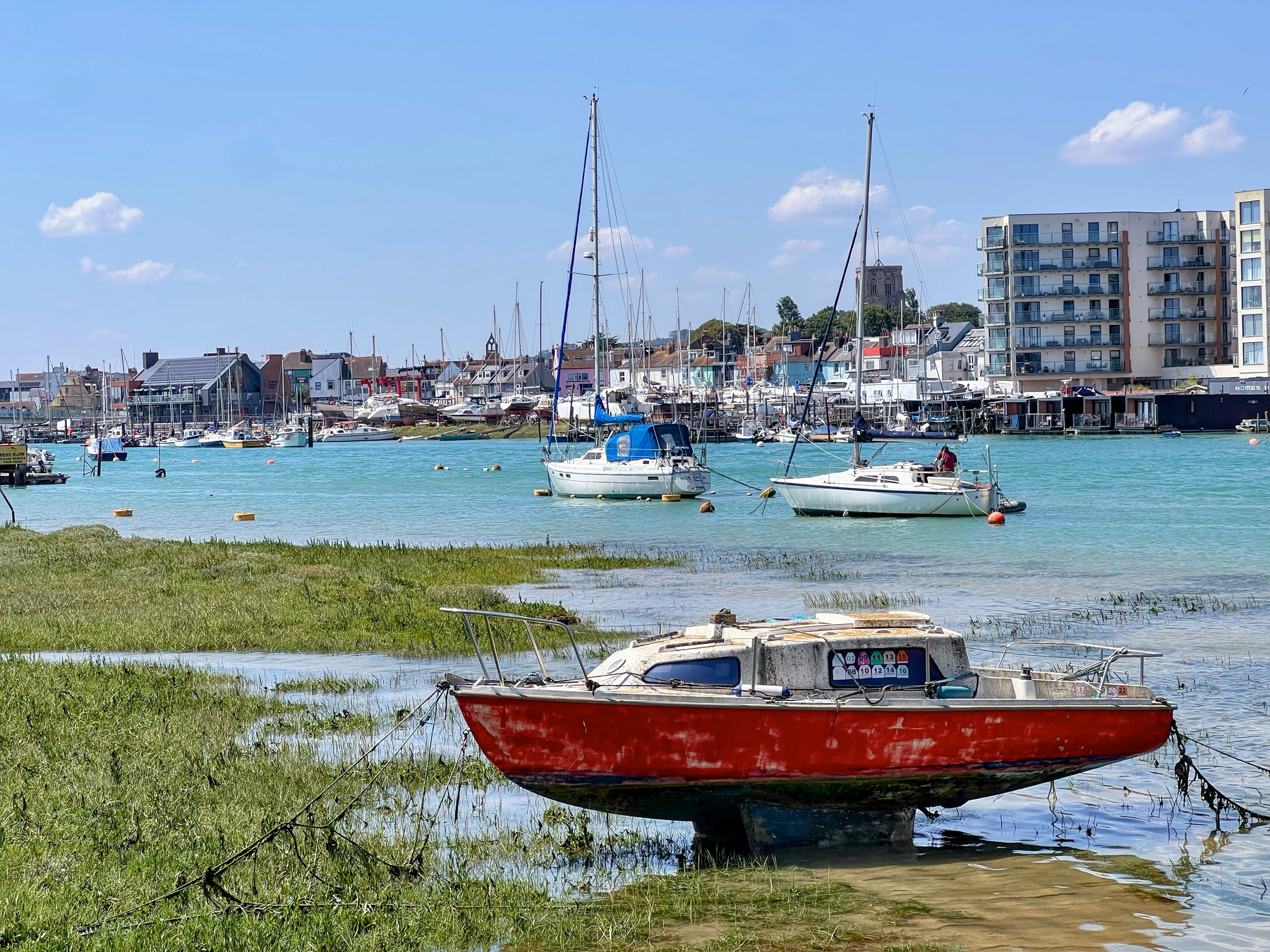 A red boat is anchored on the shore with several sailboats on the water and buildings in the background under a bright blue sky.