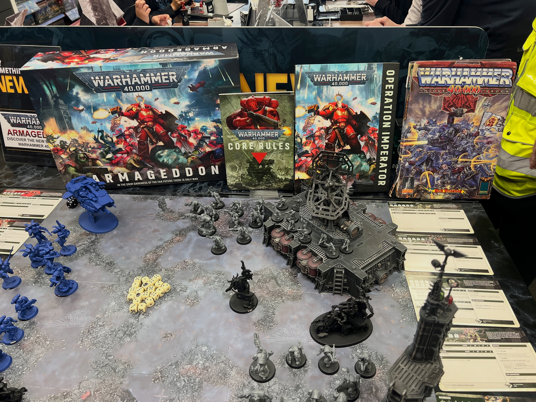 A tabletop game scene features Warhammer 40,000 miniatures, terrain, and rulebooks, including Armageddon and Operation Imperator.