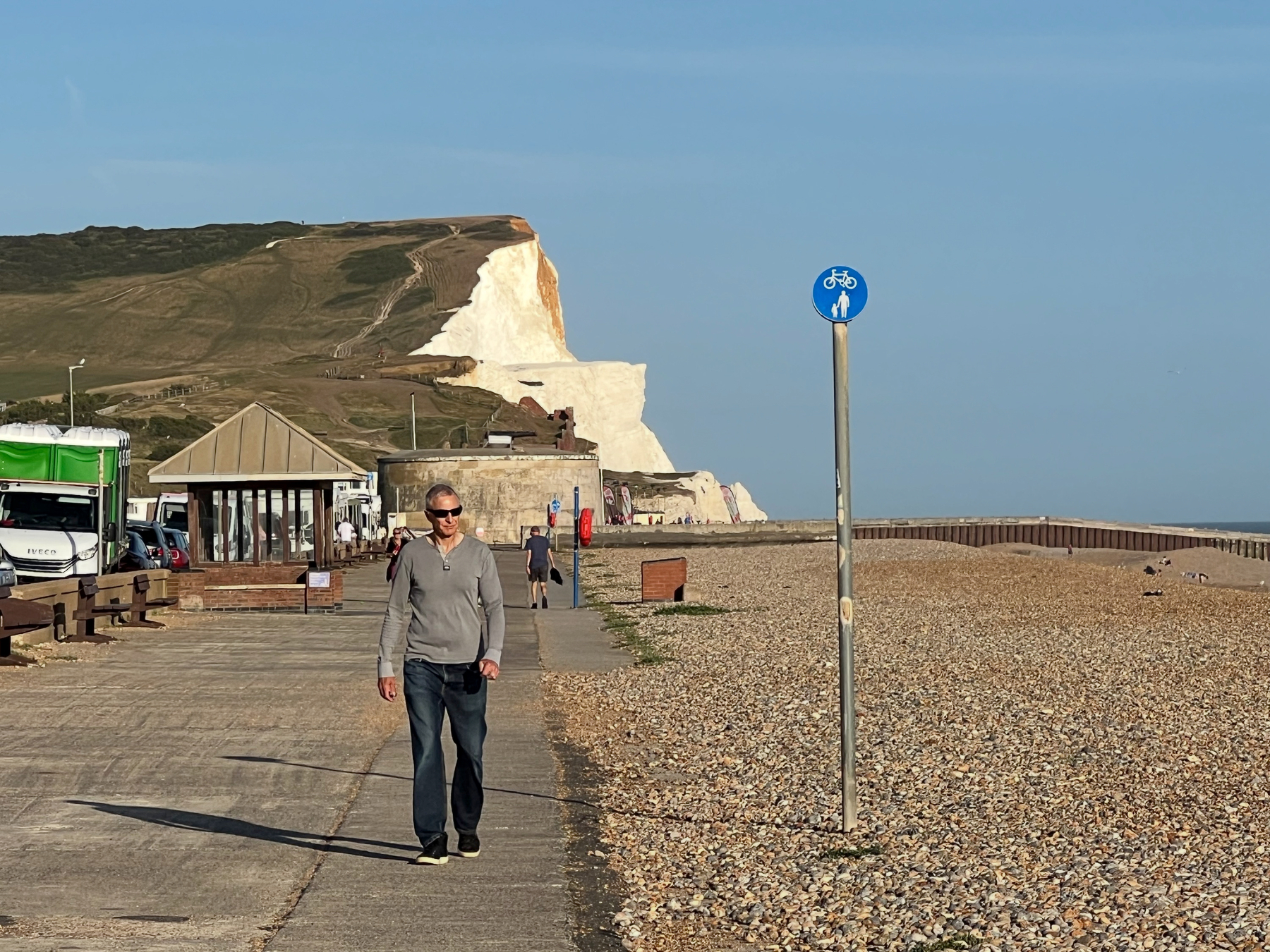 A man is walking on a paved path next to a pebble beach with white chalk cliffs in the background under a clear blue sky.