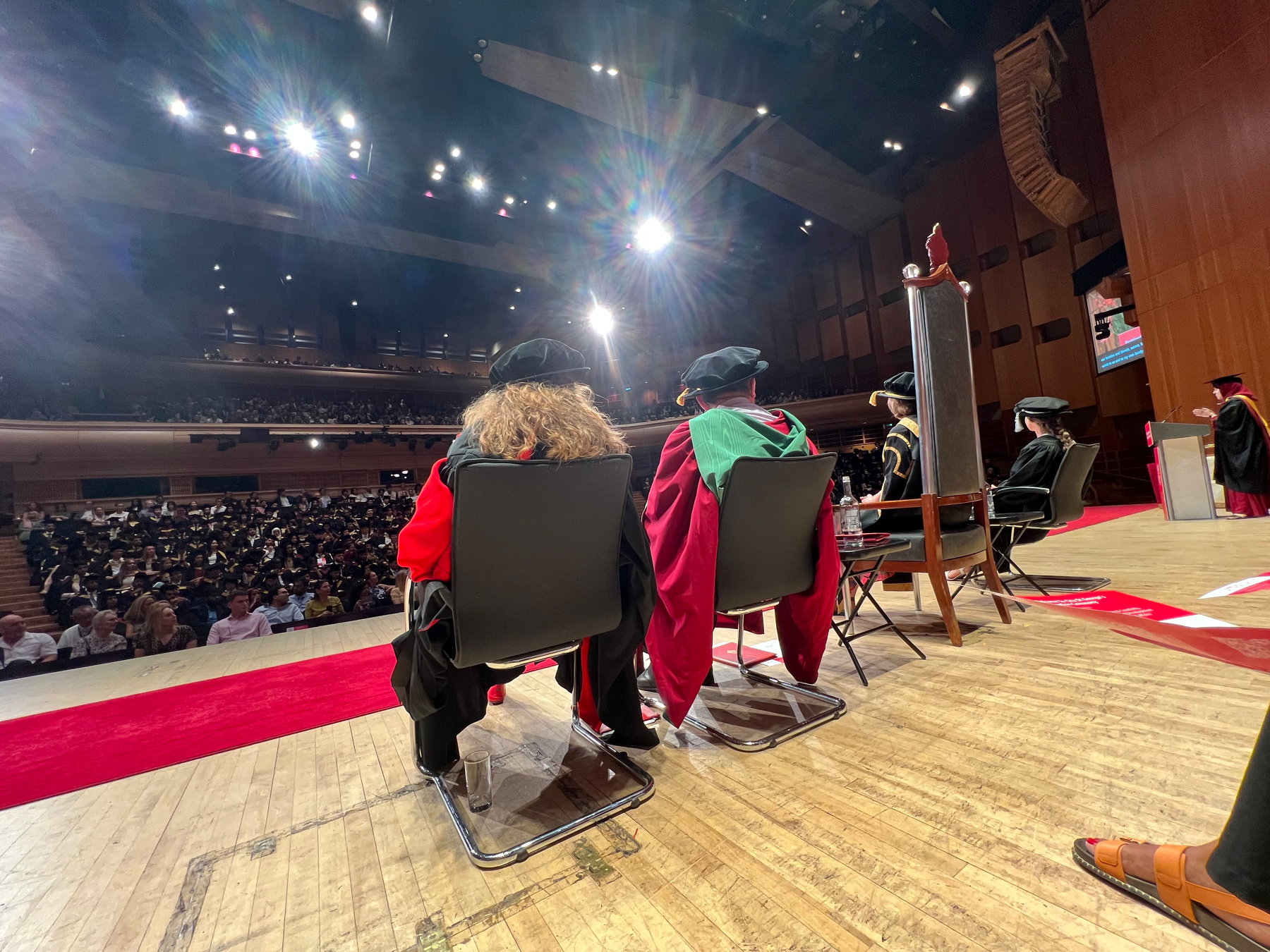 Academics in traditional graduation robes are seated on stage facing a large audience in an auditorium.