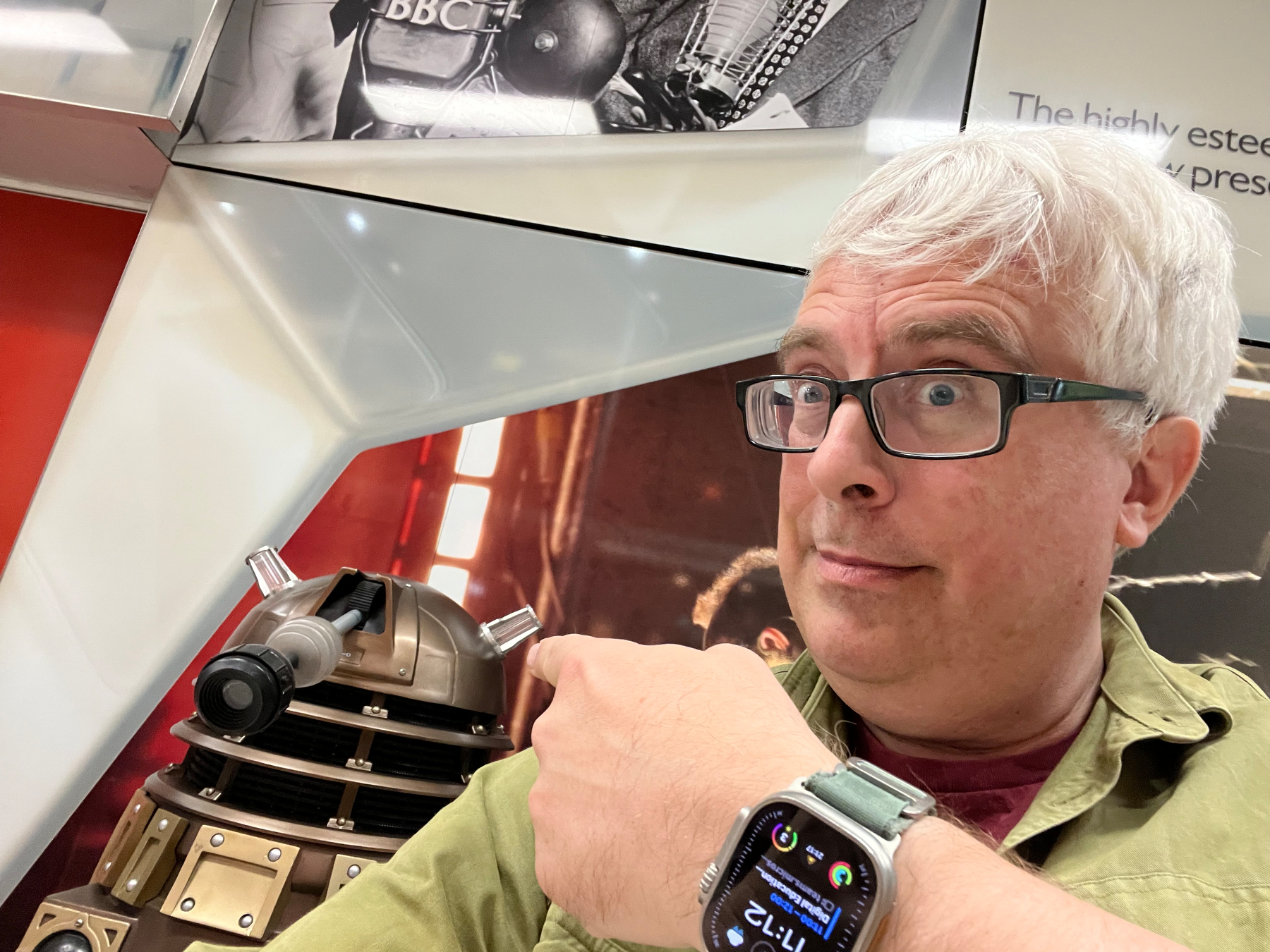 A person with glasses excitedly poses in front of a Dalek from Doctor Who.