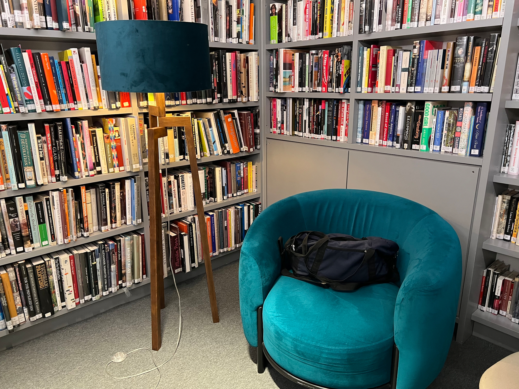 A cozy reading nook features turquoise upholstered armchair with a bag on it, surrounded by bookshelves filled with books and accompanied by a tall floor lamp with a matching turquoise shade.