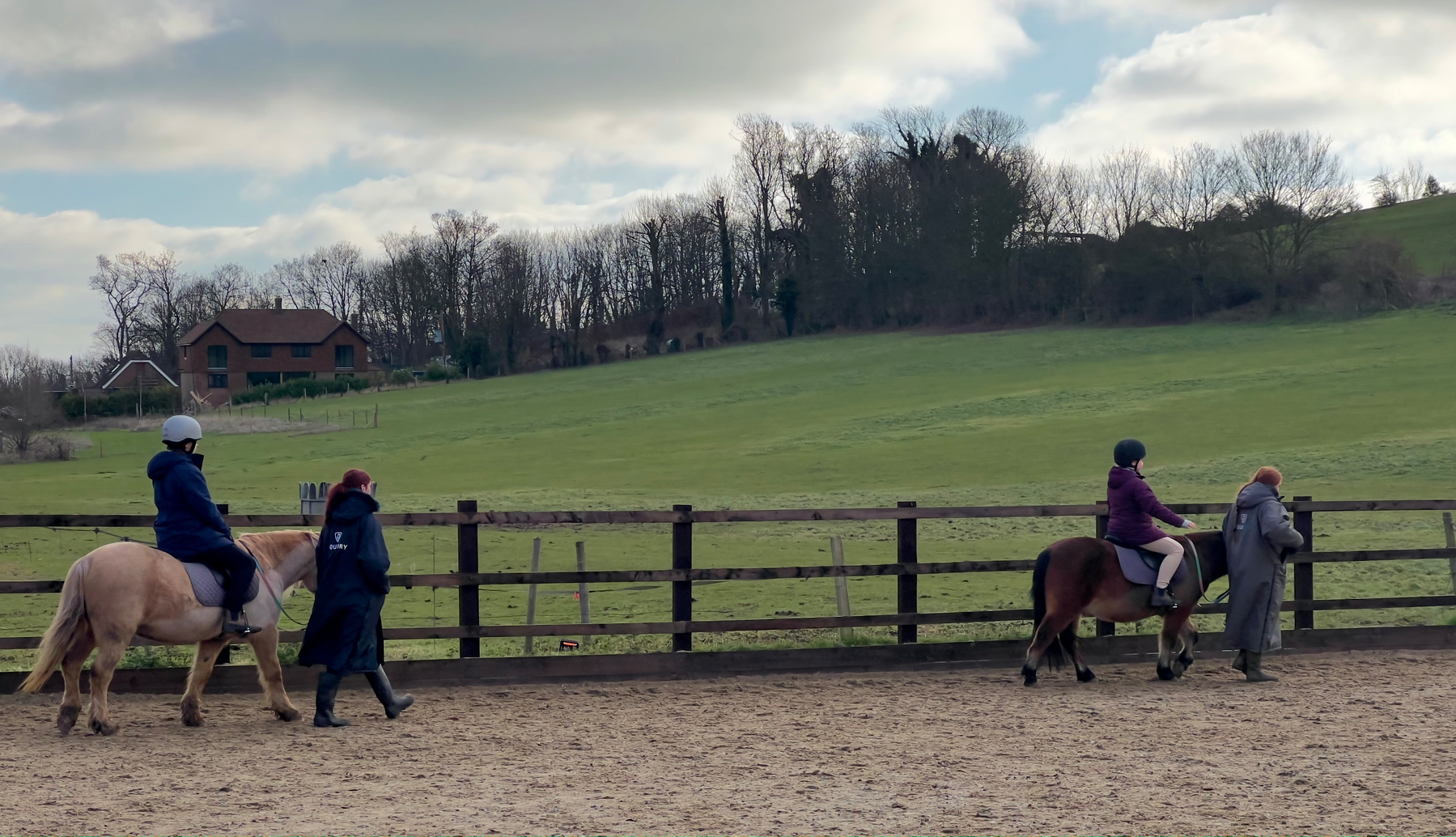 Two people are horseback riding in a fenced area, accompanied by two individuals walking beside them, with a grassy field and house in the background.
