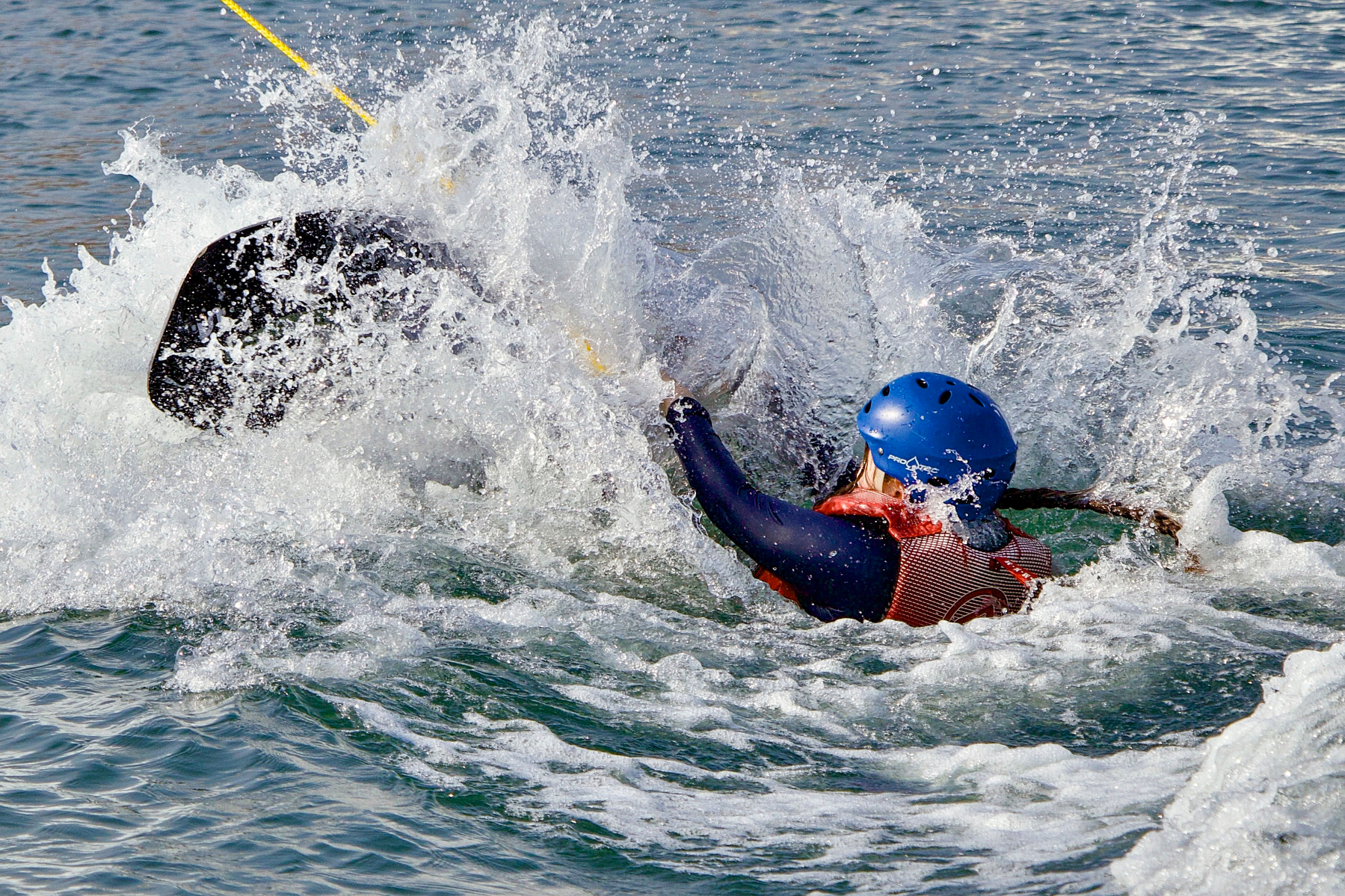 A person wearing a helmet and life vest is falling off a wakeboard into the water, creating a large splash.