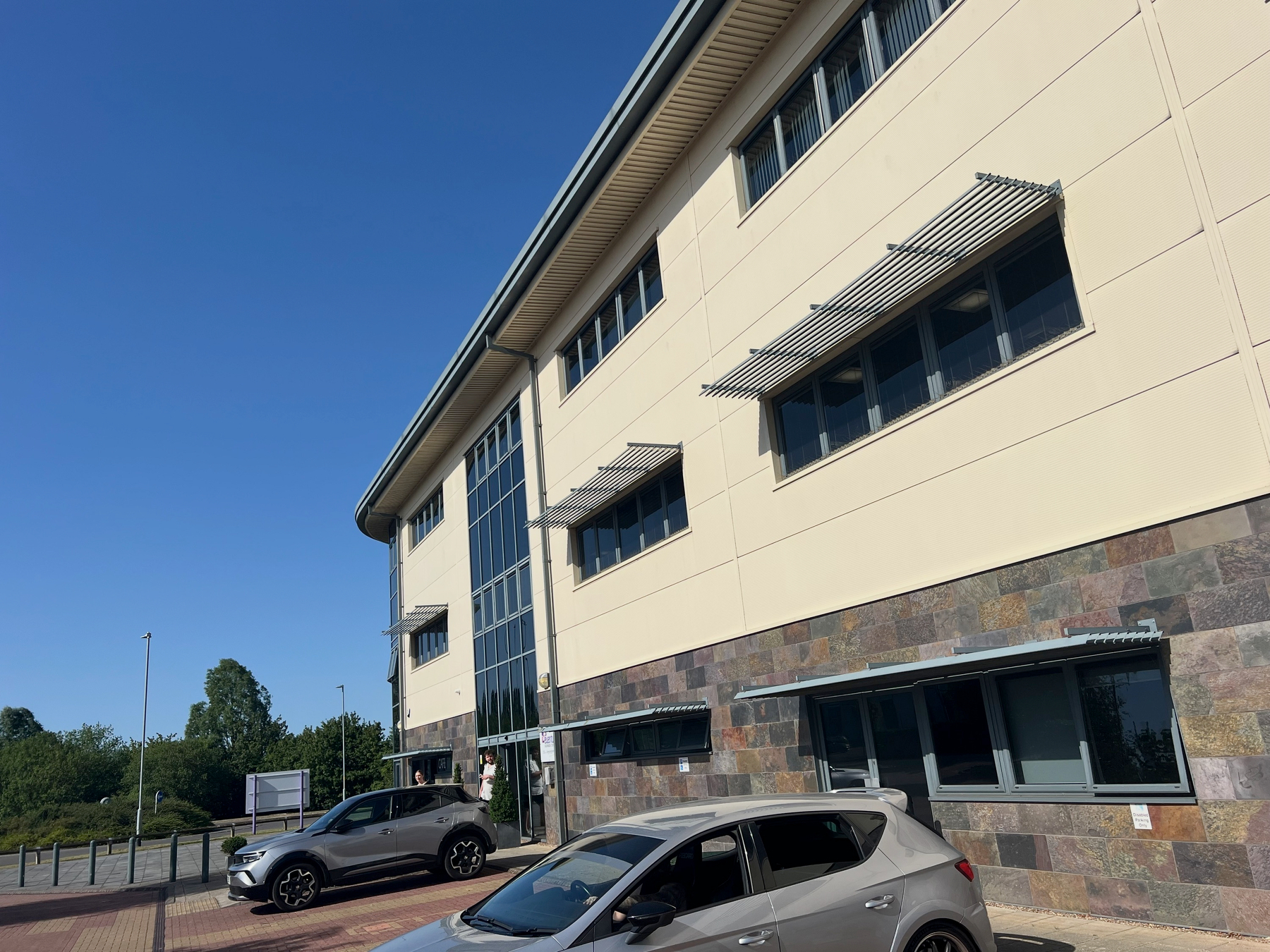 A modern office building with large windows and parked cars in front is shown under a clear blue sky.